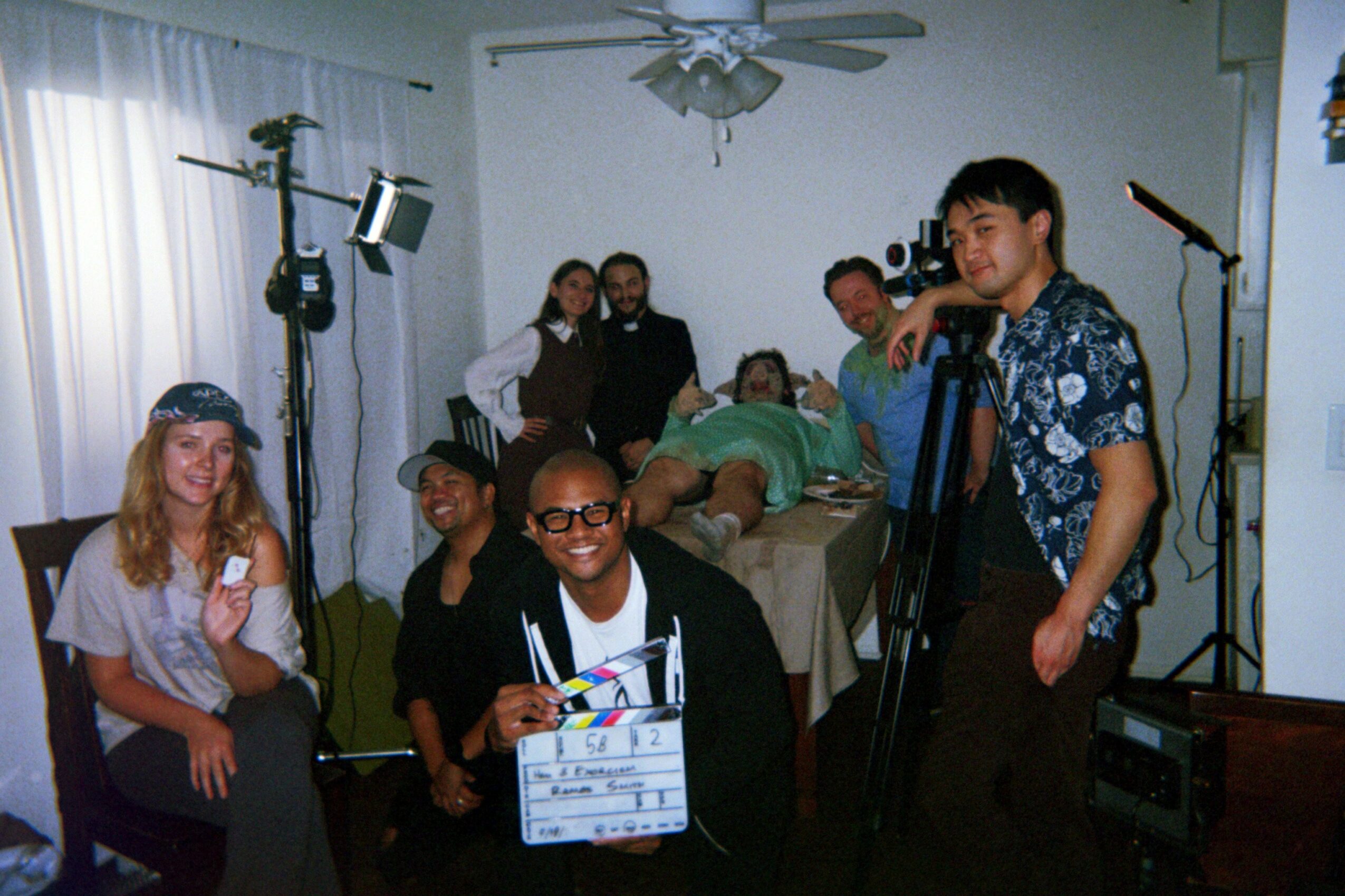 Group of people in a room, some sitting and some standing, with camera equipment and a person holding a clapperboard.