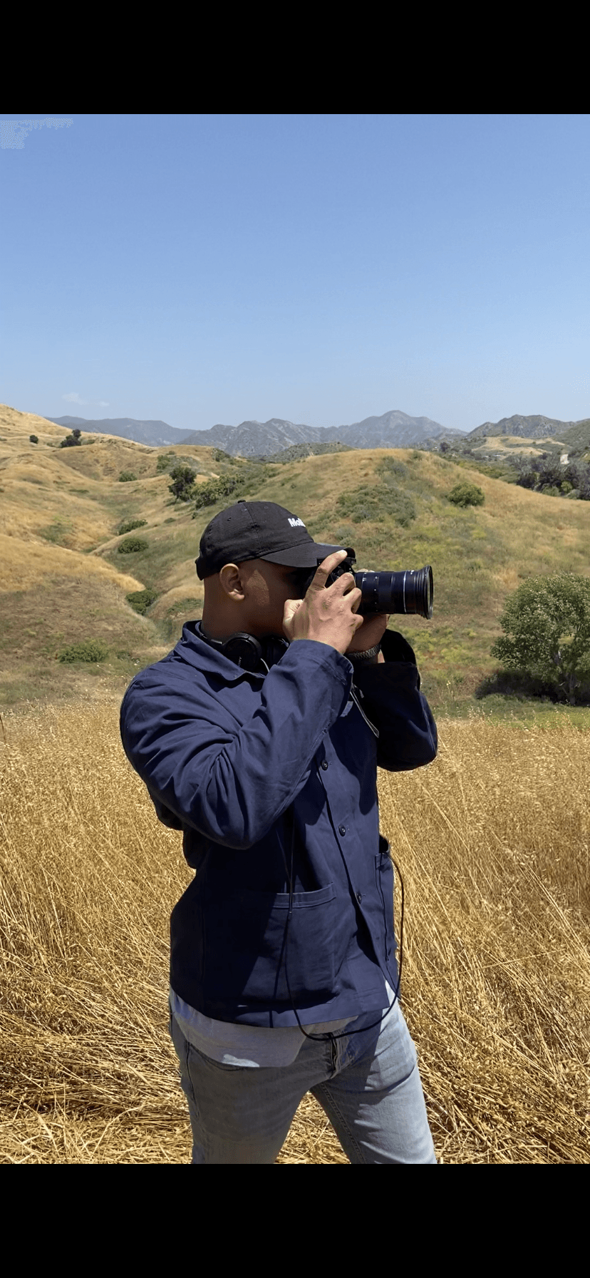 Person in dark jacket and cap holding a camera outdoors in a grassy landscape with hills and mountains, clear blue sky.