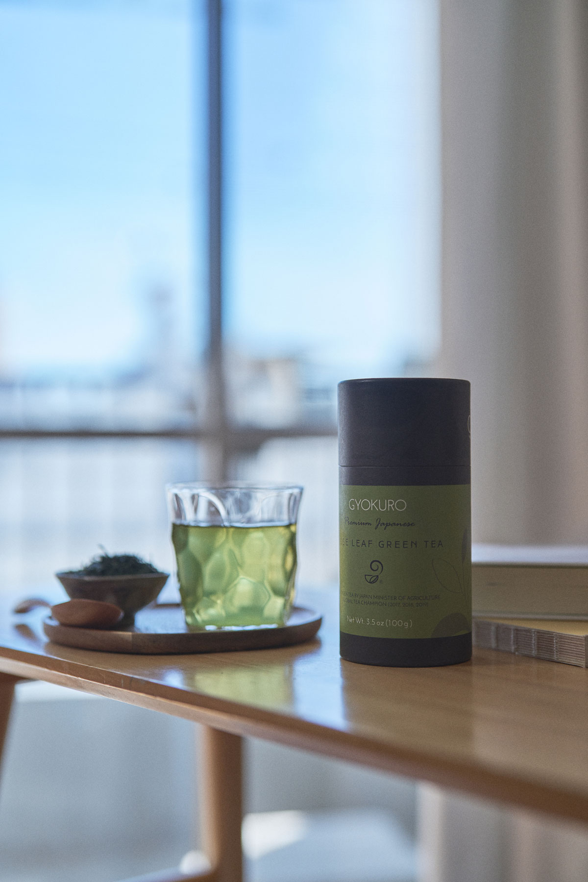 A glass of green tea, a container, and a small bowl on a wooden table near a window with a blurred cityscape background.