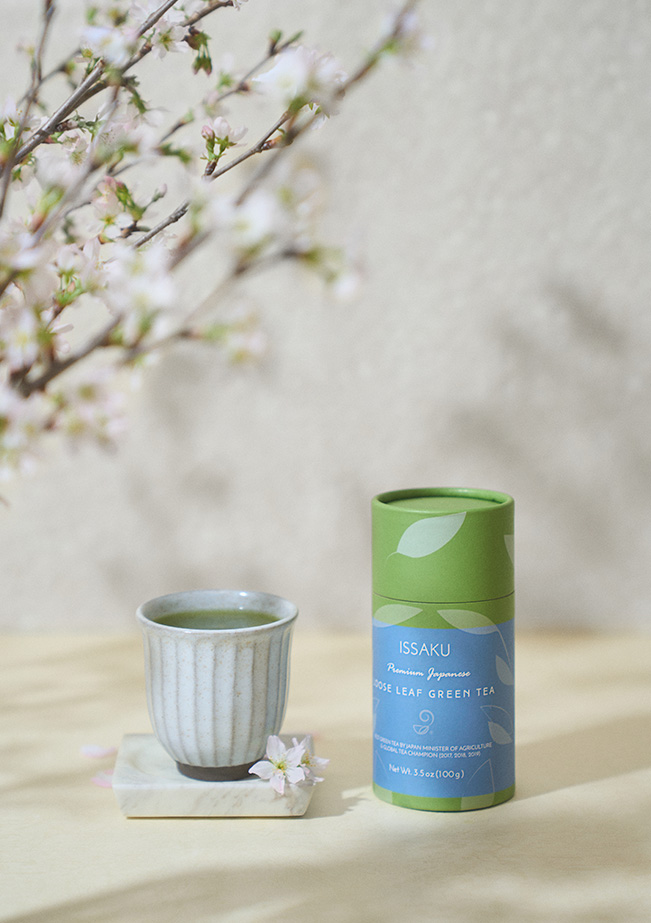 A small ceramic cup on a white surface with a green container beside it, against a light background with blurred flowers.