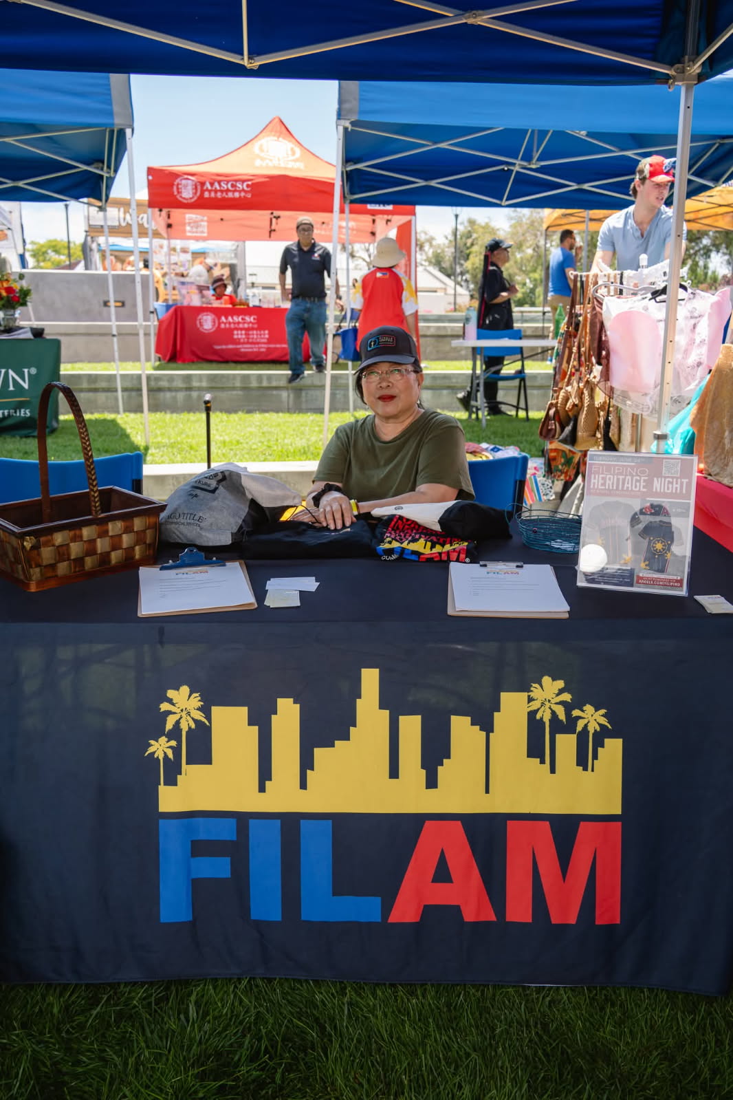Person sitting at a table with a FILAM banner, outdoor event with tents and people in background.