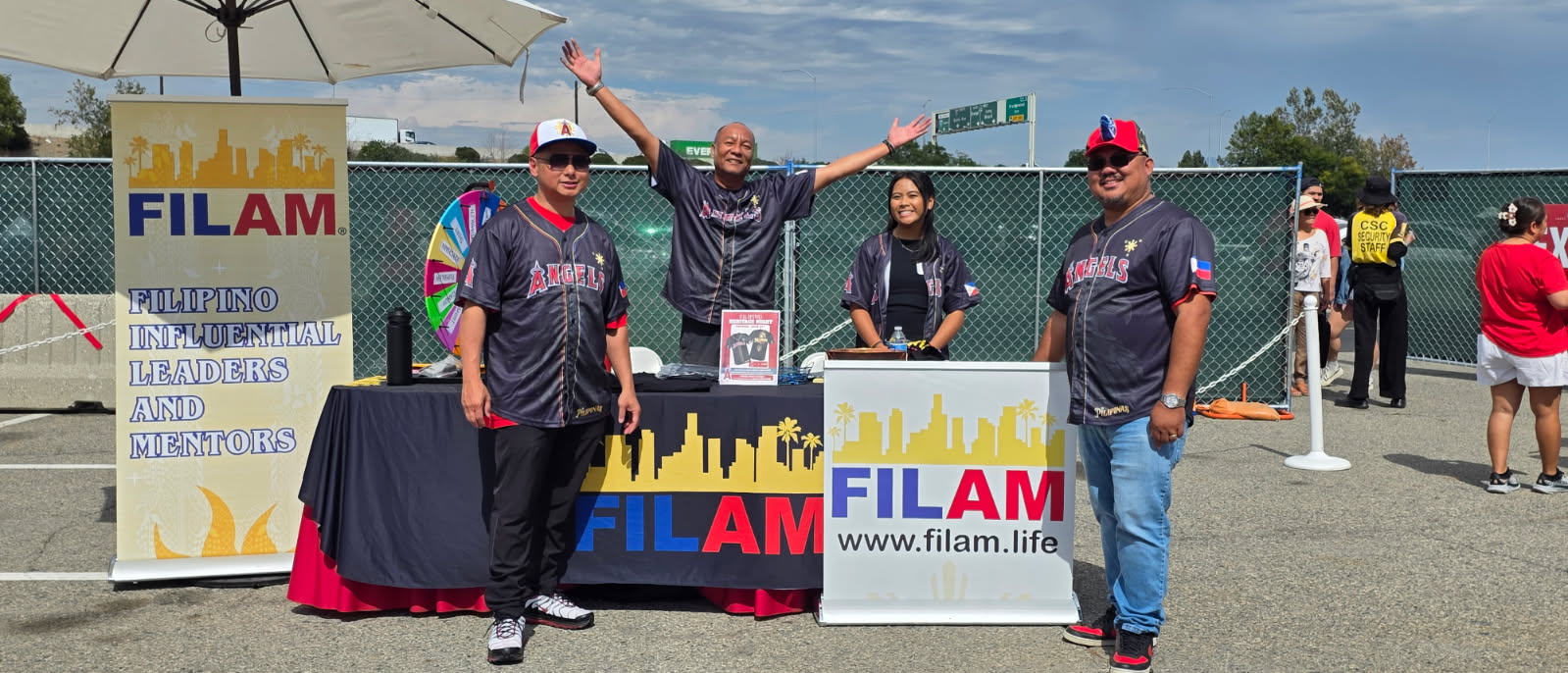 Four people stand behind a FILAM booth at an outdoor event, with a table, banner, and umbrella, under a blue sky.