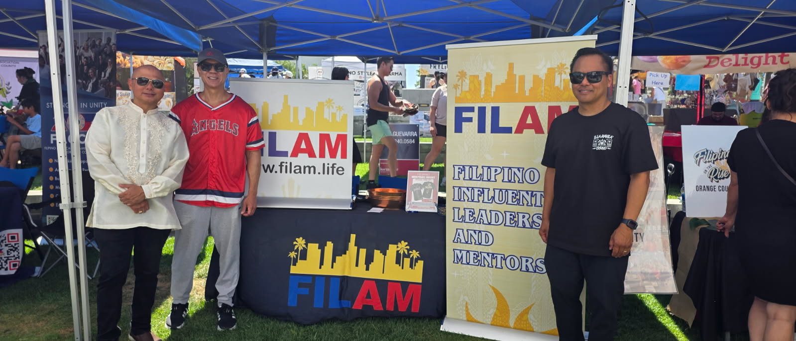 Three people standing outdoors at a booth with banners and signs, under a blue canopy, in a grassy area.