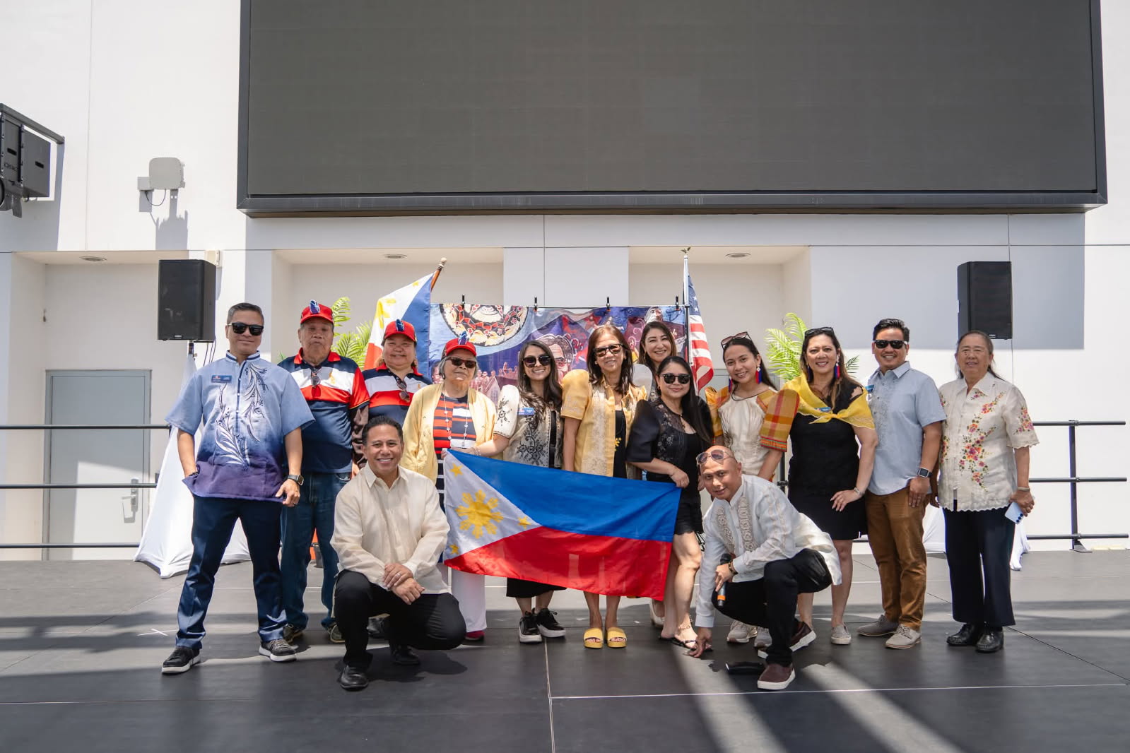 Group of people holding flags and banners, standing outdoors in front of a building, some wearing sunglasses, smiling.