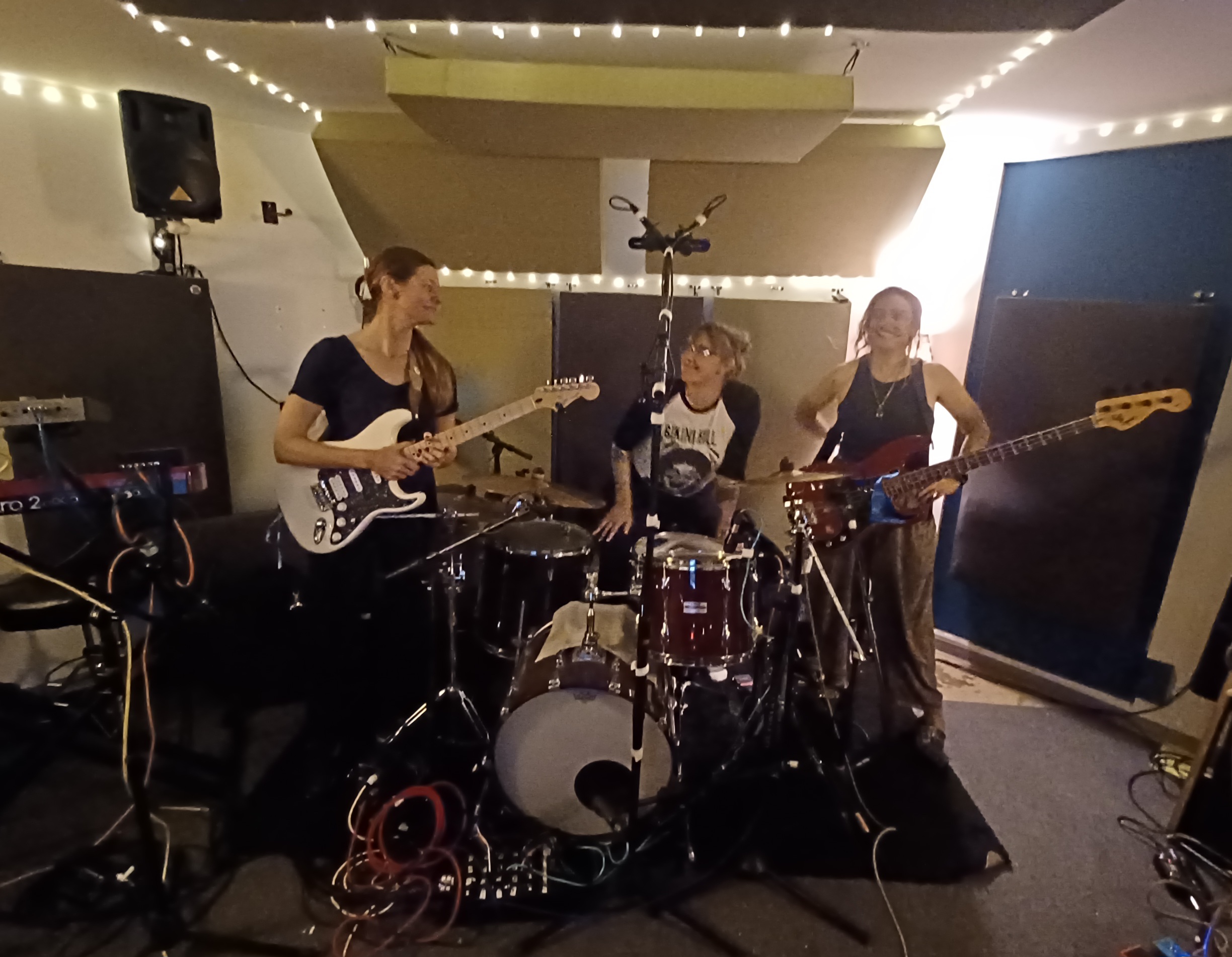 Three women playing guitars and drums in a band, with equipment and stage lighting, in an indoor setting.
