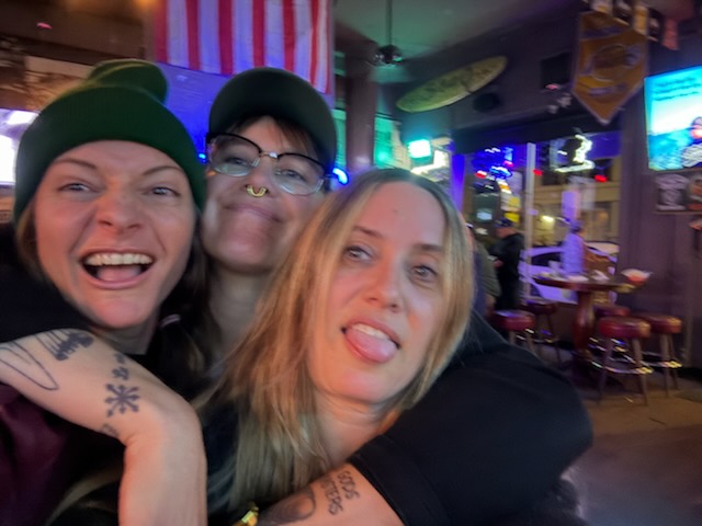 Three people smiling and sticking out their tongues in a lively indoor setting with colorful lights.