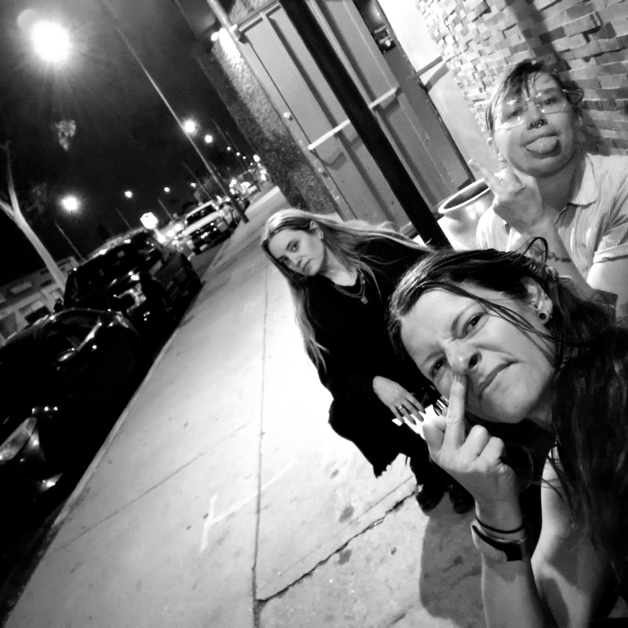 Three women taking a selfie on a city sidewalk at night, with parked cars and streetlights in the background.