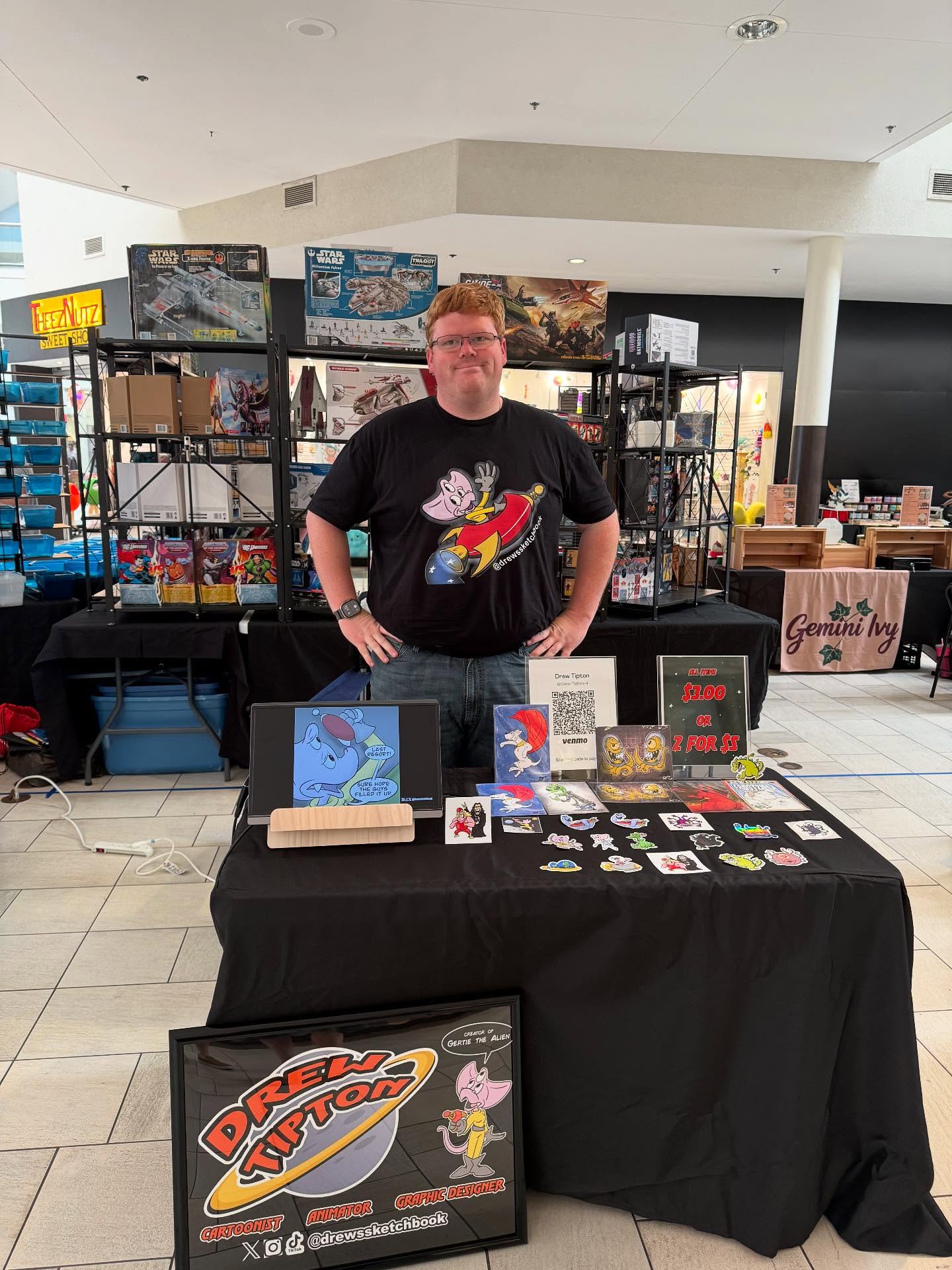 Person standing behind a table with comic books and merchandise, in a shopping mall setting.