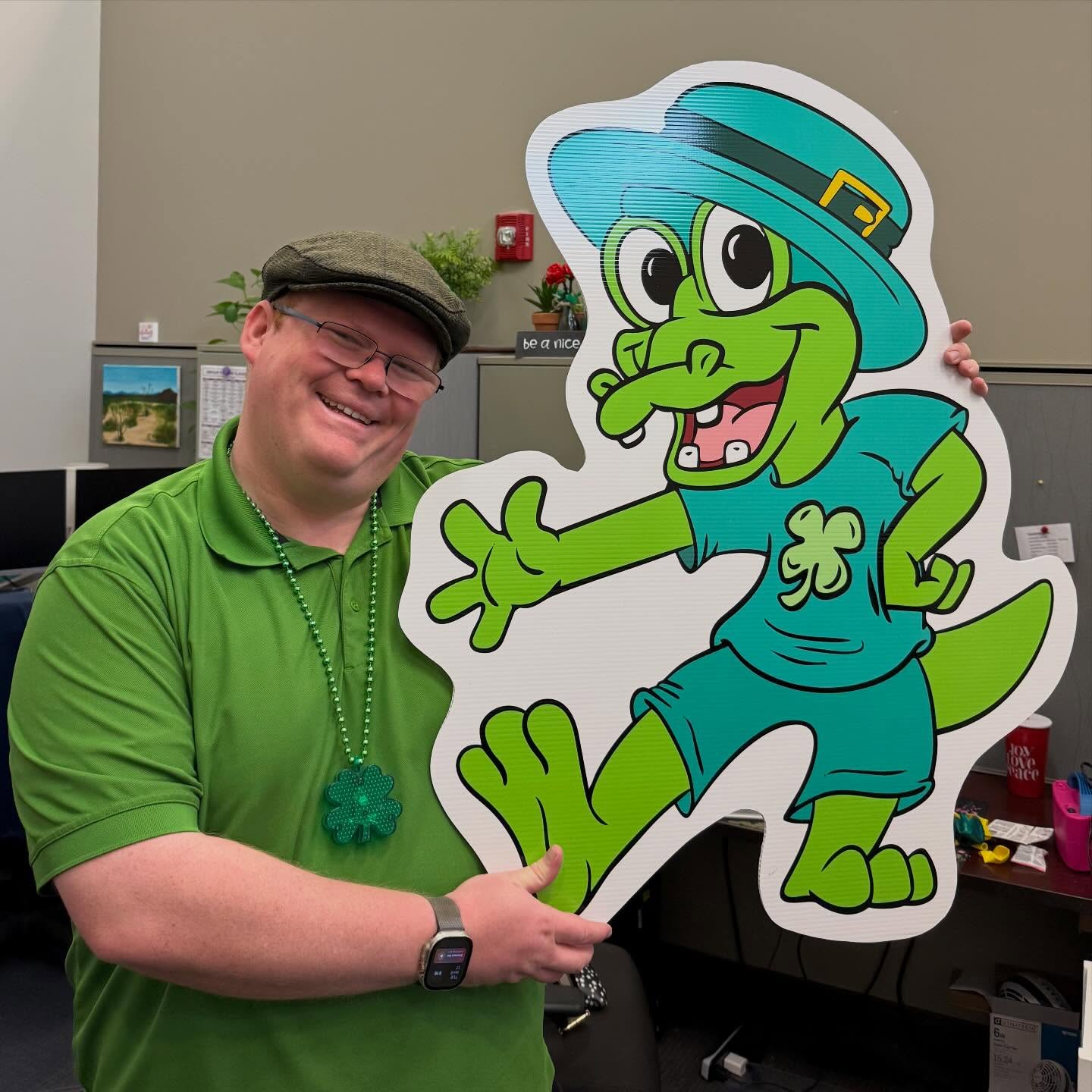 Man in green shirt and hat smiling holding a large cartoon leprechaun cutout in an office.