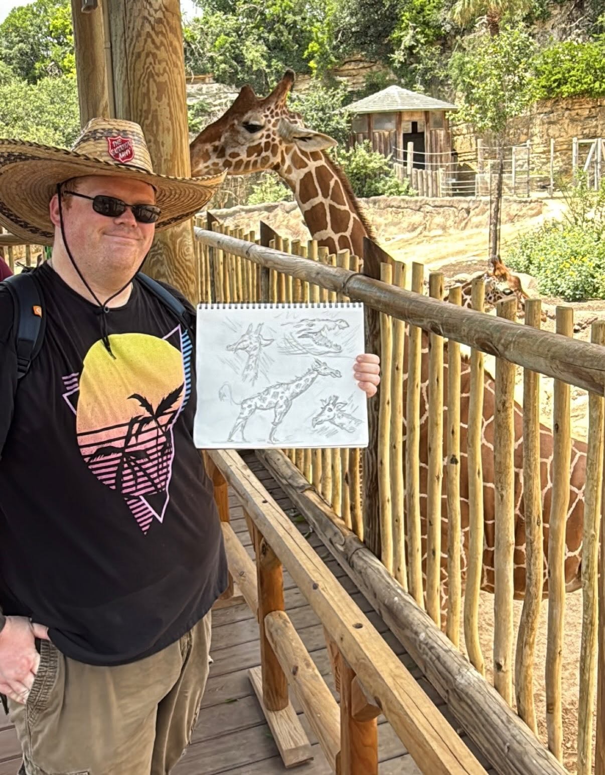 Person wearing a sombrero and sunglasses holding a drawing of a giraffe at zoo, with giraffes in enclosure behind.