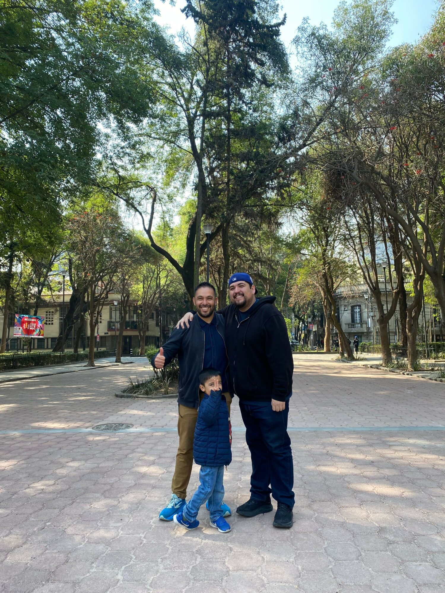 Two people standing outdoors on a paved path with trees and buildings in the background.