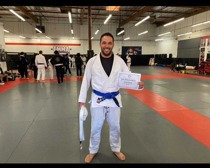Man in a white martial arts gi with a blue belt holding a certificate, smiling in a gym with people in the background.