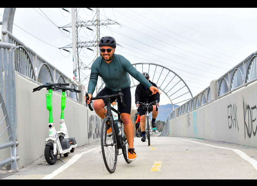 Person riding a bicycle on a bridge with a scooter parked nearby, other cyclists in the background, overcast sky.