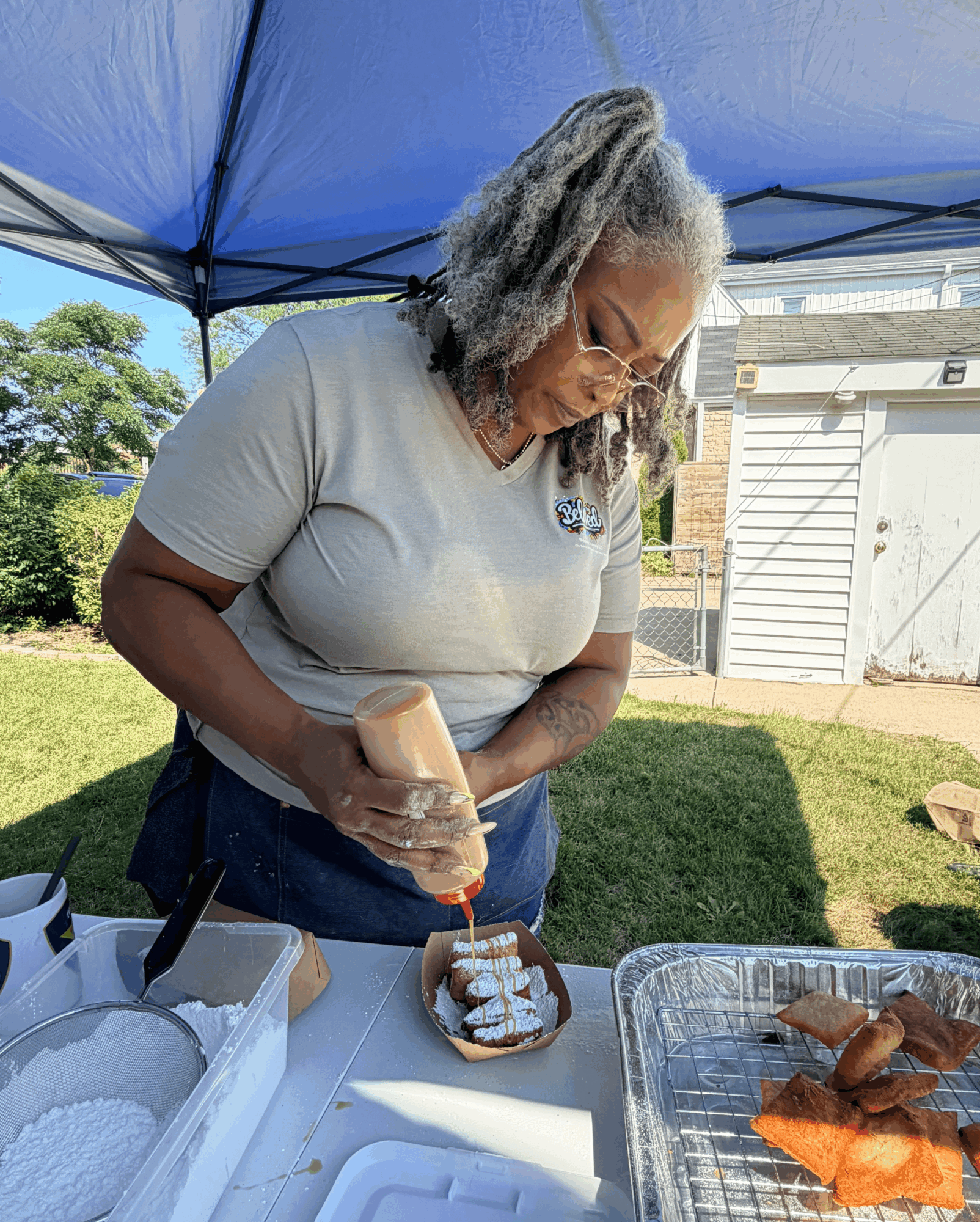 Woman with gray hair and glasses pours sauce on a dessert, outdoors under a blue canopy, with food on table.