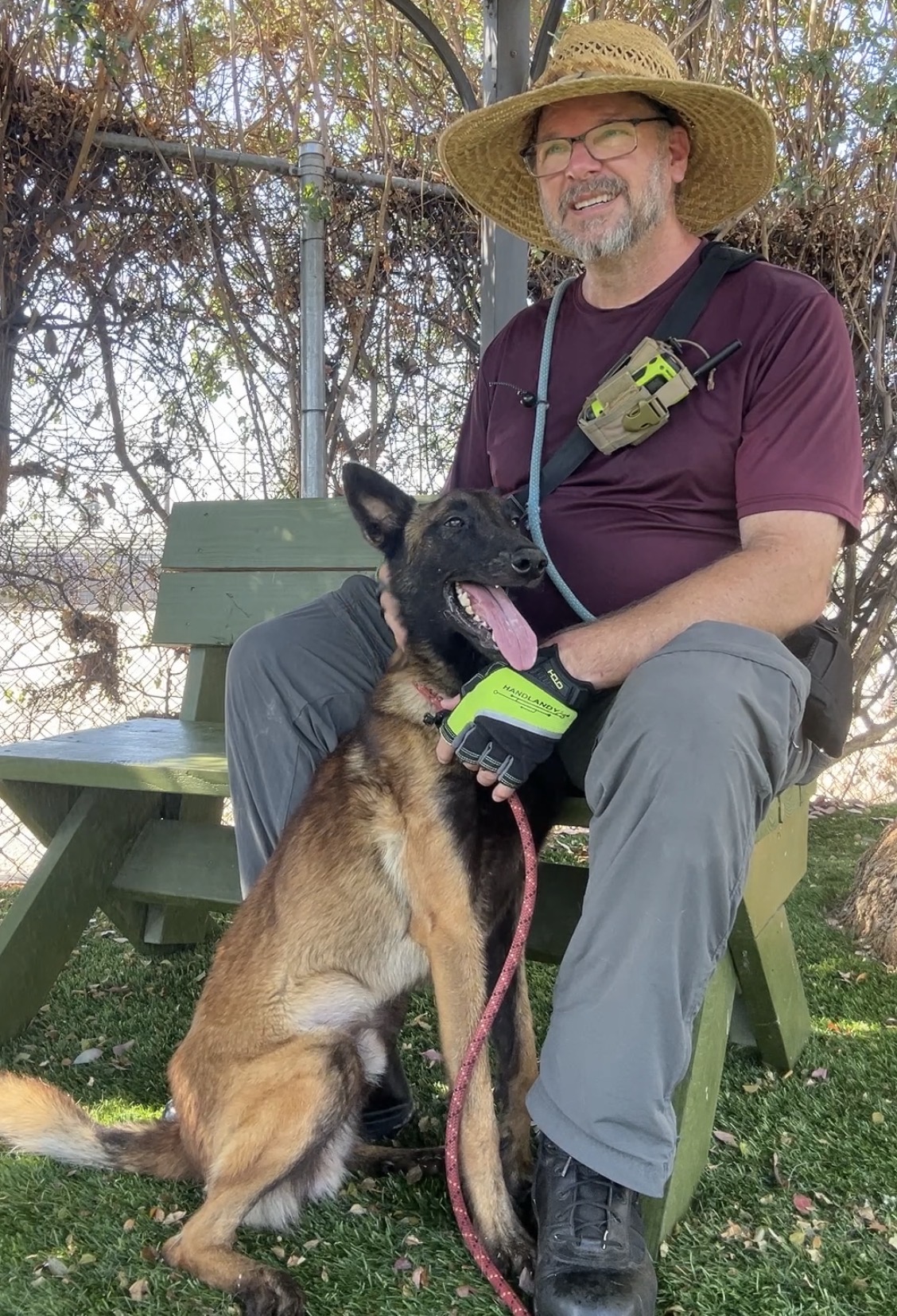 Man with glasses and a straw hat sitting on a bench, holding a Belgian Malinois dog on a leash outdoors.