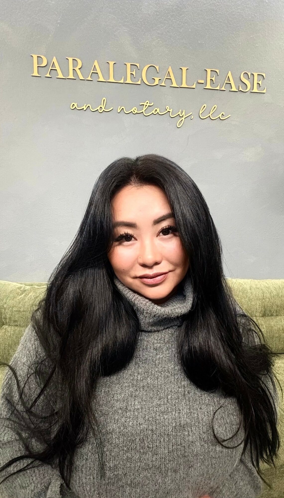 Young woman with long dark hair wearing a gray turtleneck sweater, smiling, sitting in front of a wall with gold text.