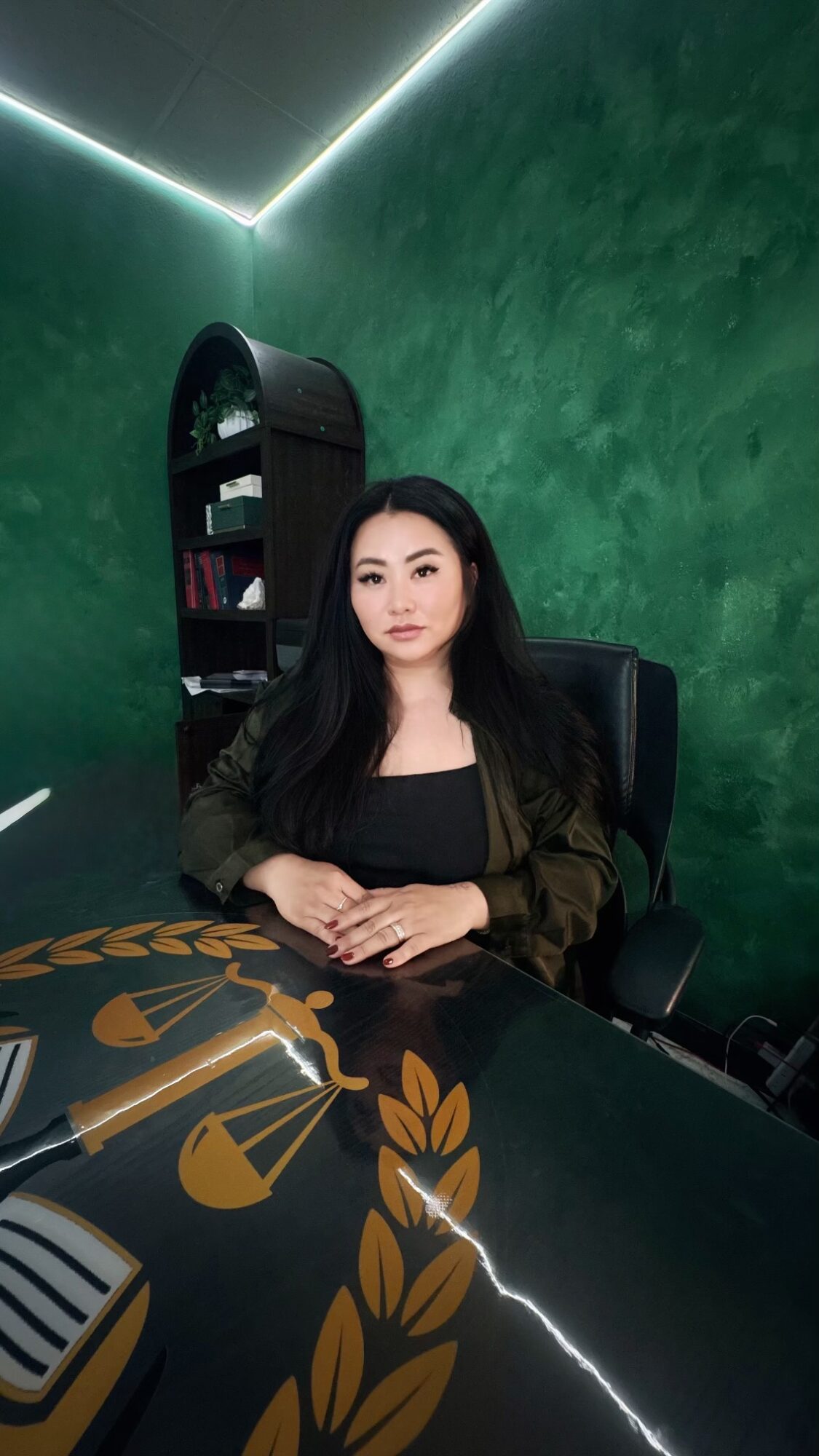 Woman with long dark hair sitting at a desk in a room with green textured wall and black furniture.