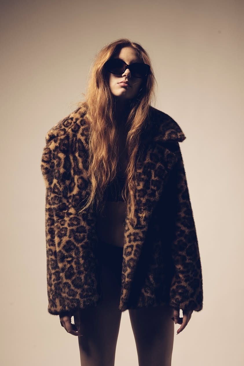 Woman with long hair wearing sunglasses and a leopard print coat, standing against a plain background.