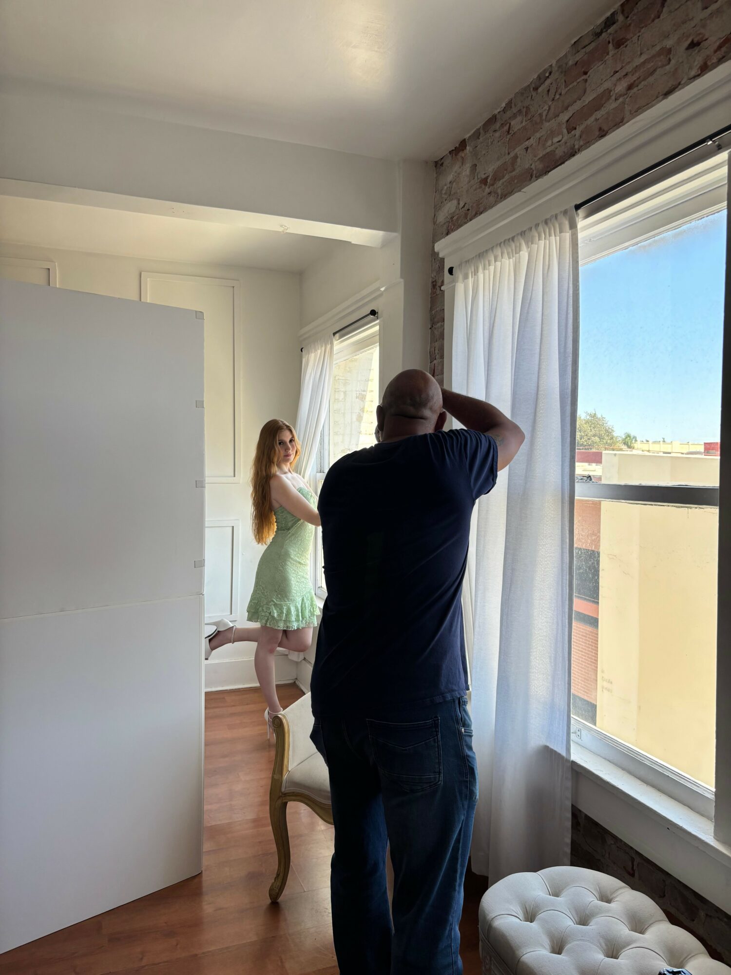 Person standing near a large window with sunlight, facing sideways, with a woman sitting on a ledge inside the room.