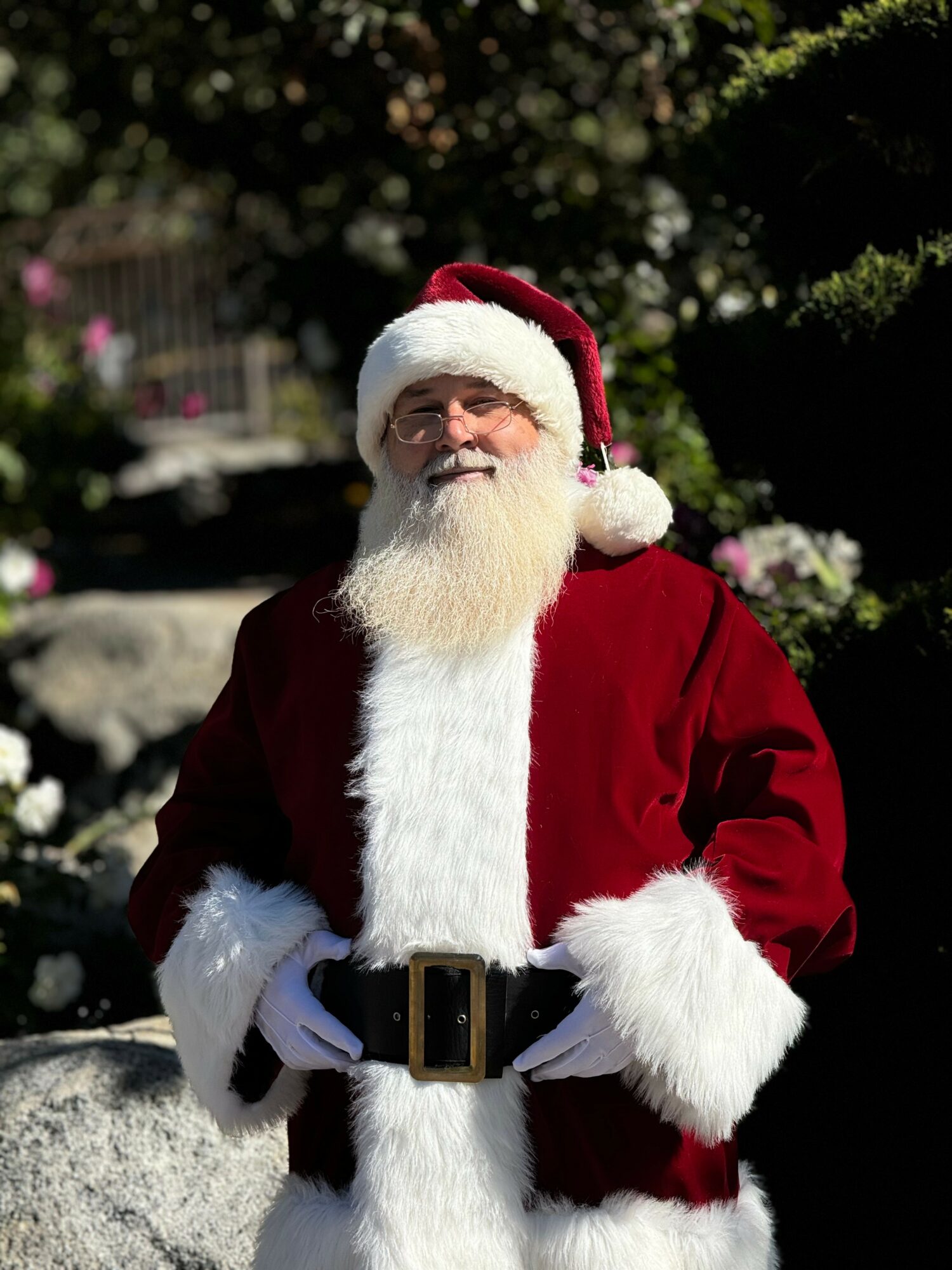 Santa Claus outdoors with a white beard, red suit, and black belt, smiling in sunlight with greenery in background.