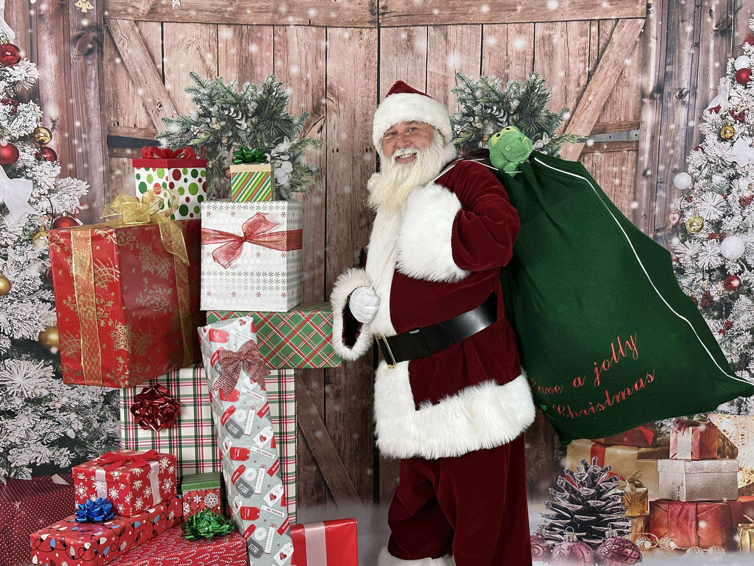 Santa Claus standing next to wrapped Christmas presents and decorated trees, holding a large green sack, in front of a wooden background.