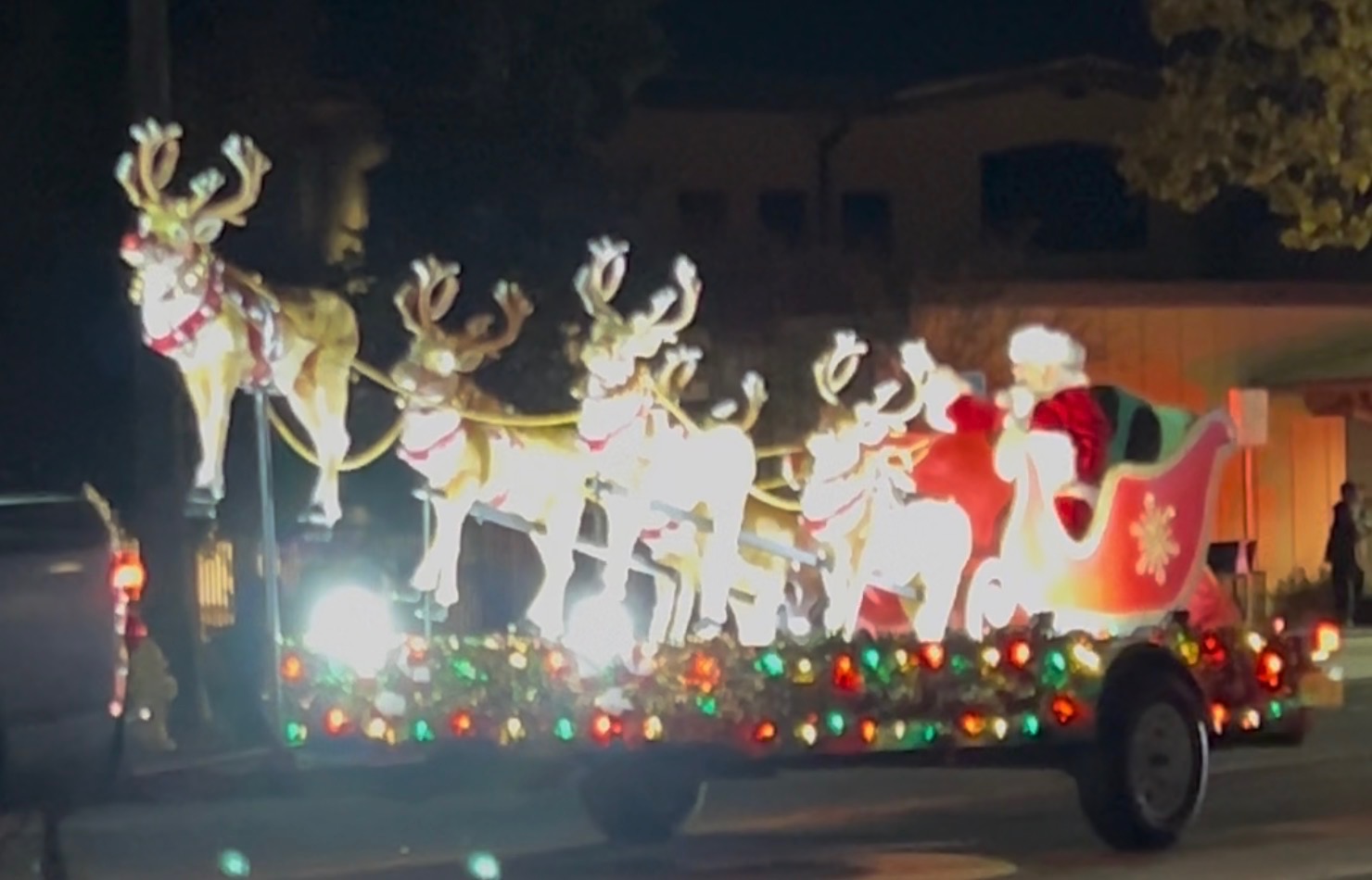 A decorated vehicle with reindeer figures and Santa Claus, illuminated with colorful lights, during a nighttime parade or event.