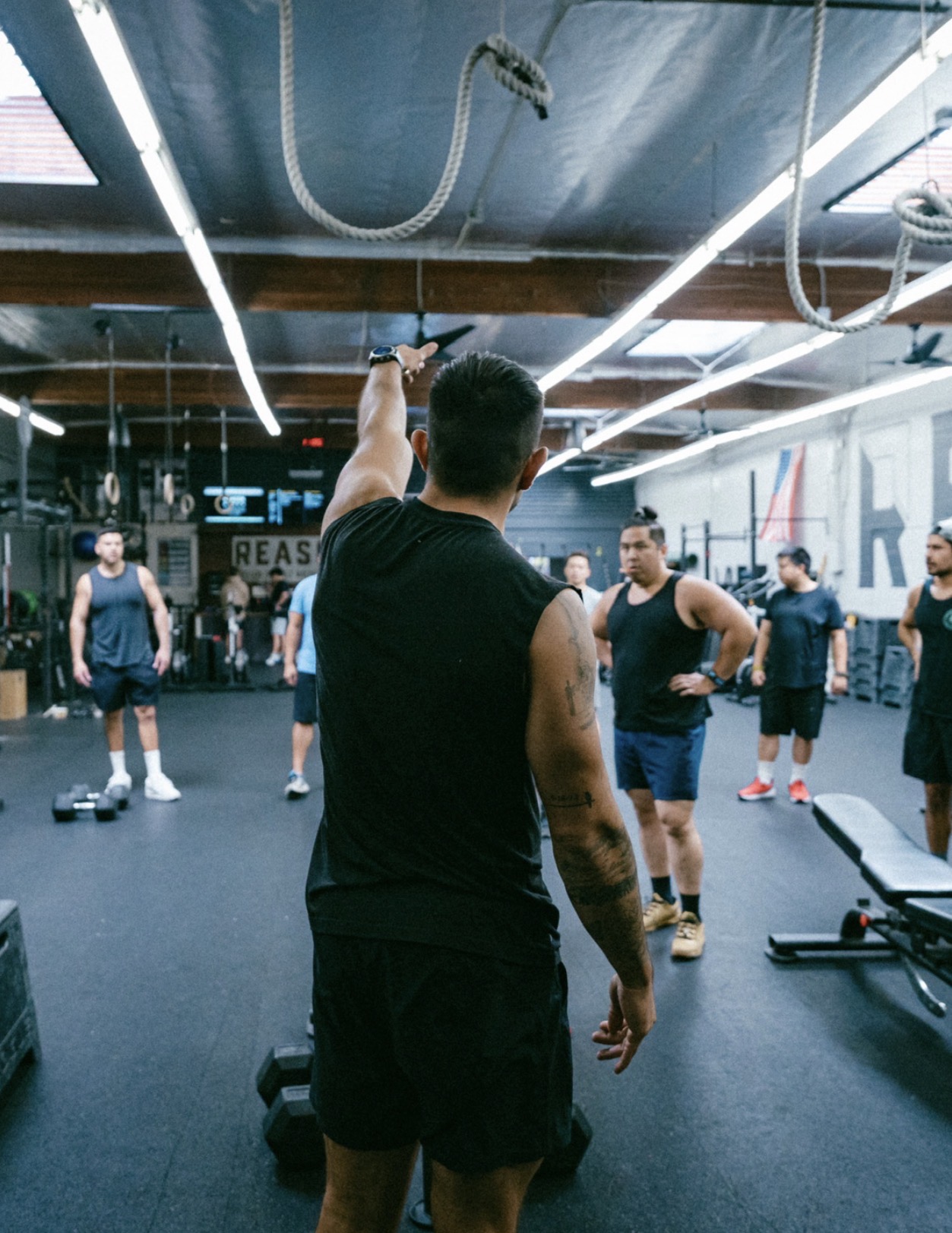 Group of people in a gym, with a person in the foreground holding a kettlebell, facing away from the camera.