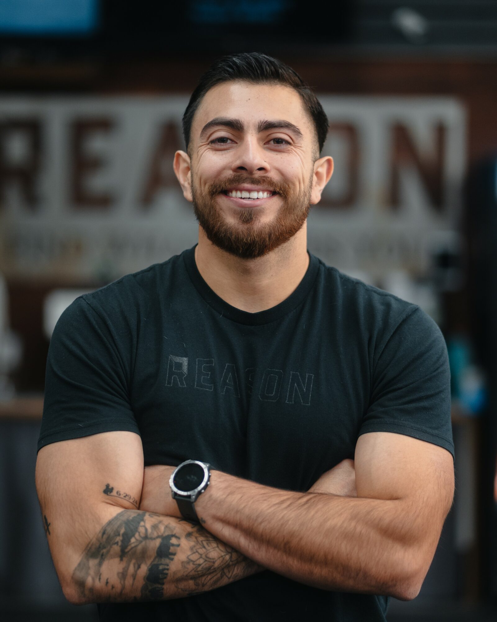Smiling man with dark hair and beard, arms crossed, wearing a black T-shirt and watch, standing outdoors.