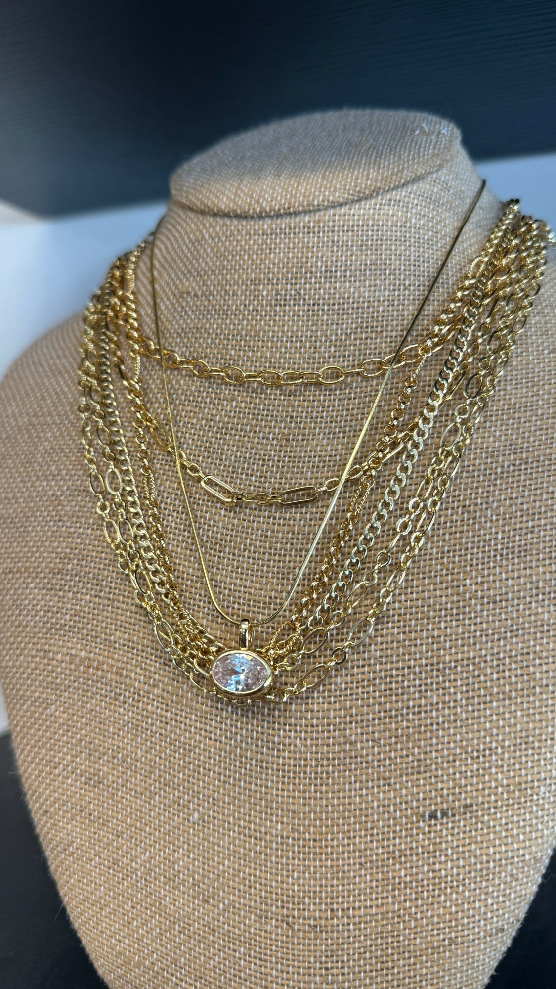 Beige jewelry display bust with multiple layered gold and silver necklaces, one with a large central pendant, against a dark background.