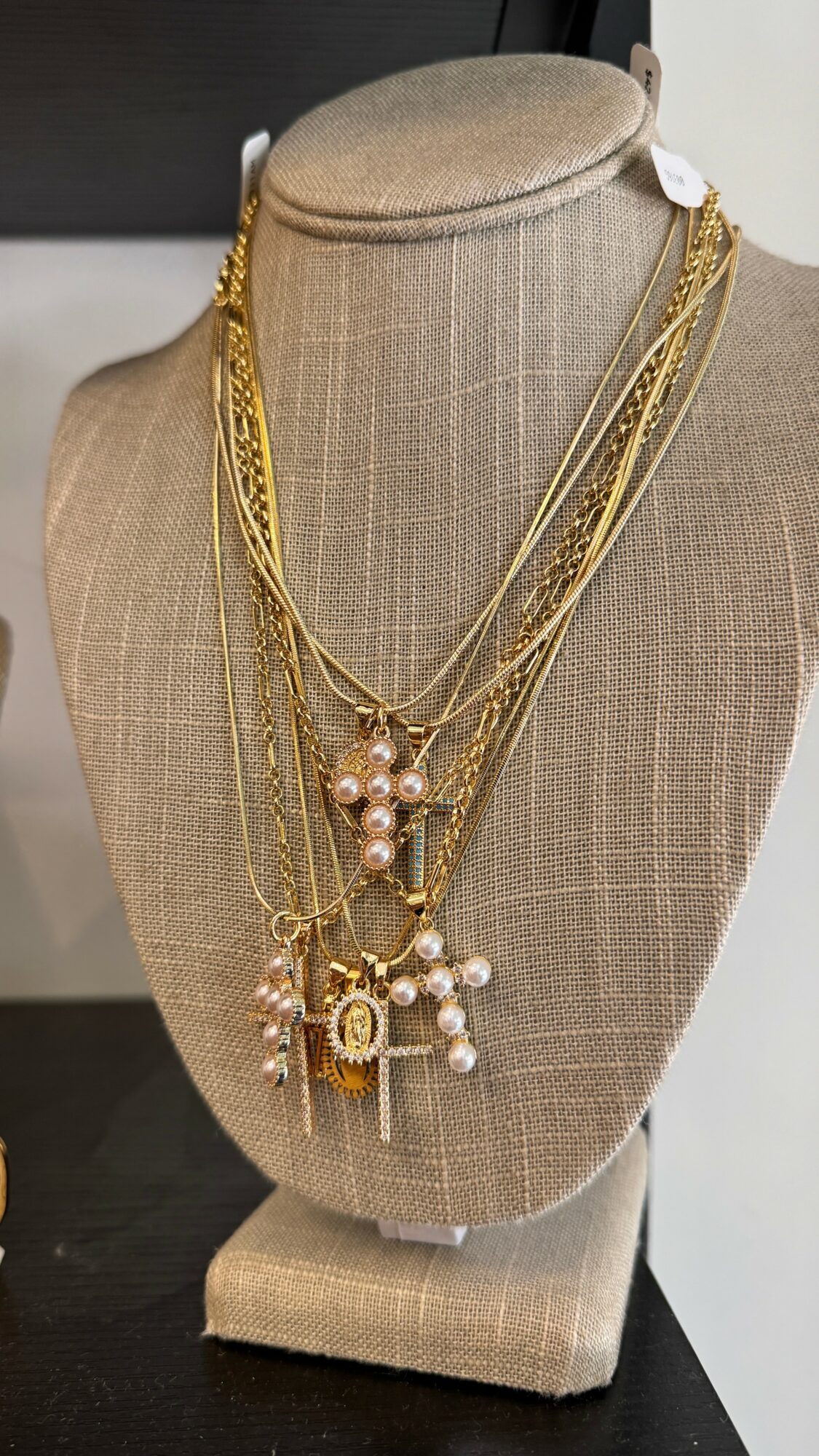 Display bust with layered gold necklaces and pearl accents, on beige fabric stand, against dark background.