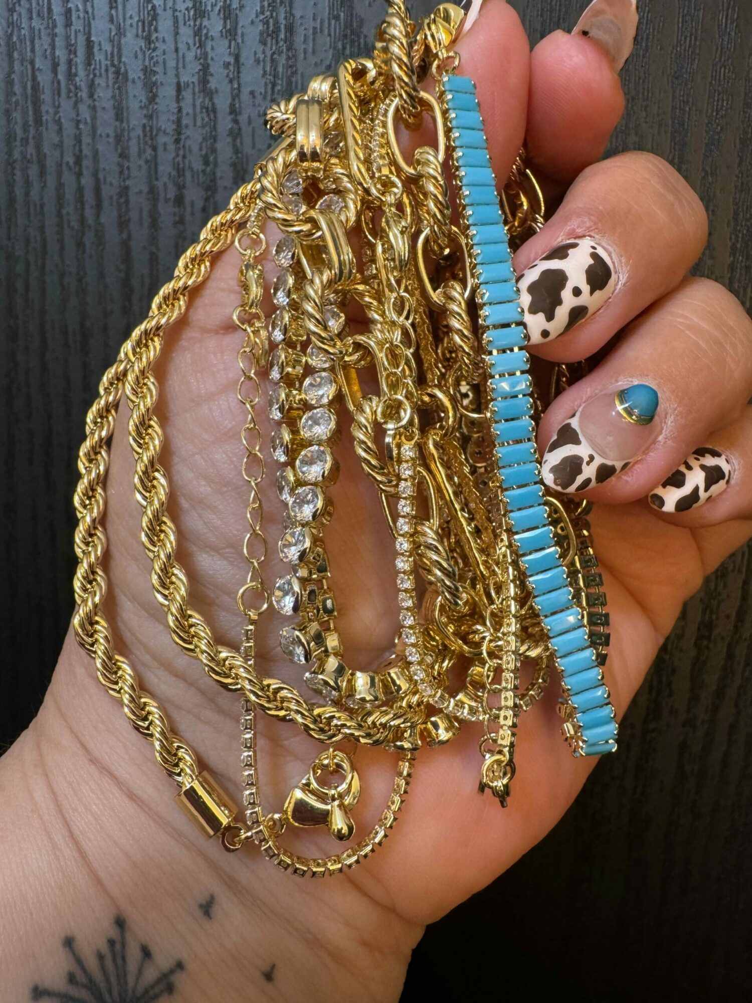 Hand with decorated nails holding multiple gold and turquoise jewelry pieces, including chains and a bracelet, on a dark surface.