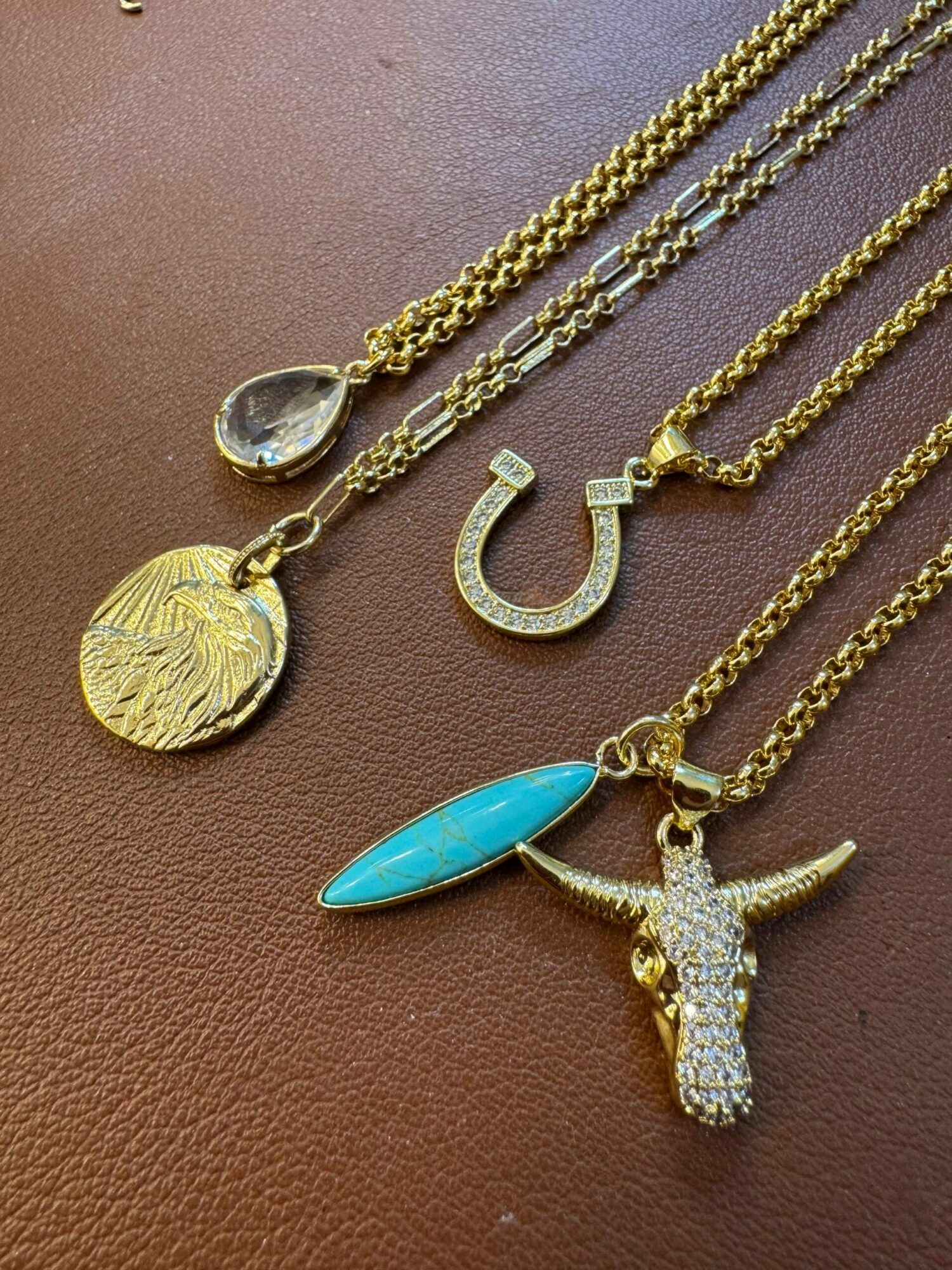 Collection of gold and silver necklaces with various pendants, including a horseshoe, turquoise stone, and textured coin, on a brown surface.