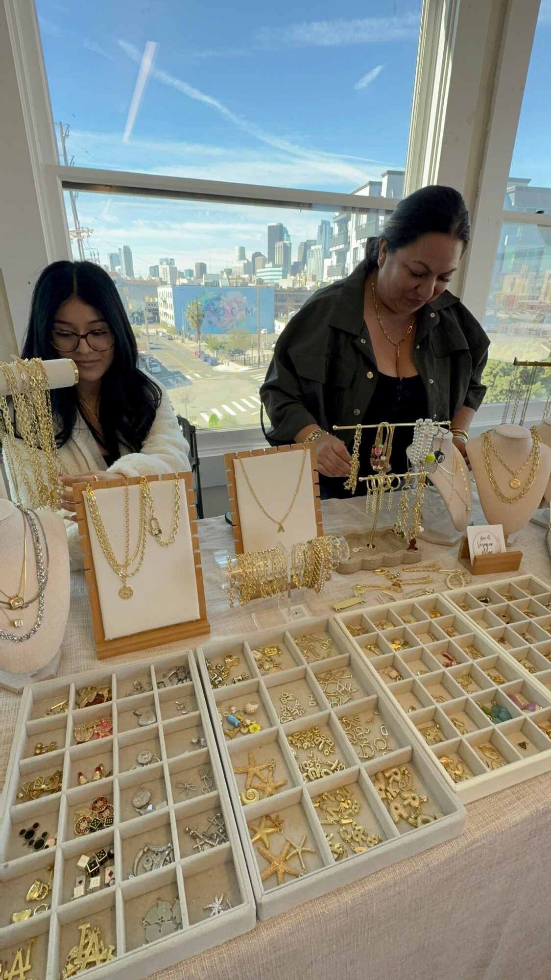 Two women browsing jewelry display with city skyline and blue sky through large window behind them.