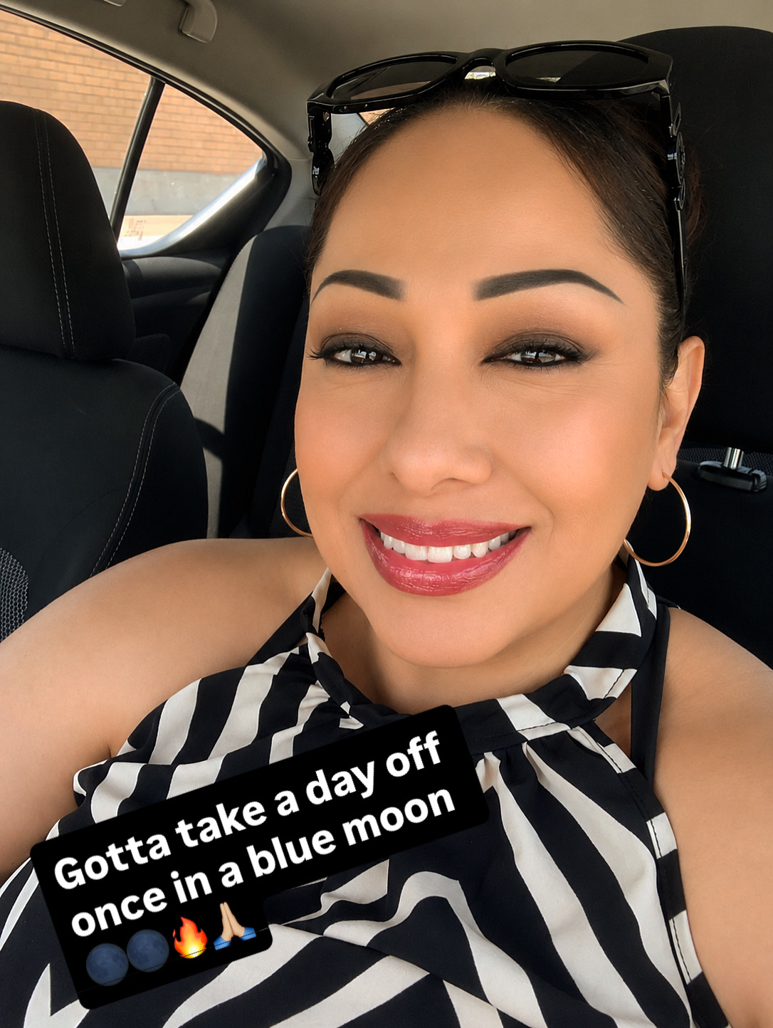 Smiling woman with sunglasses on her head, wearing hoop earrings and a striped top, sitting in a car. Caption: 'Gotta take a day off once in a blue moon.'