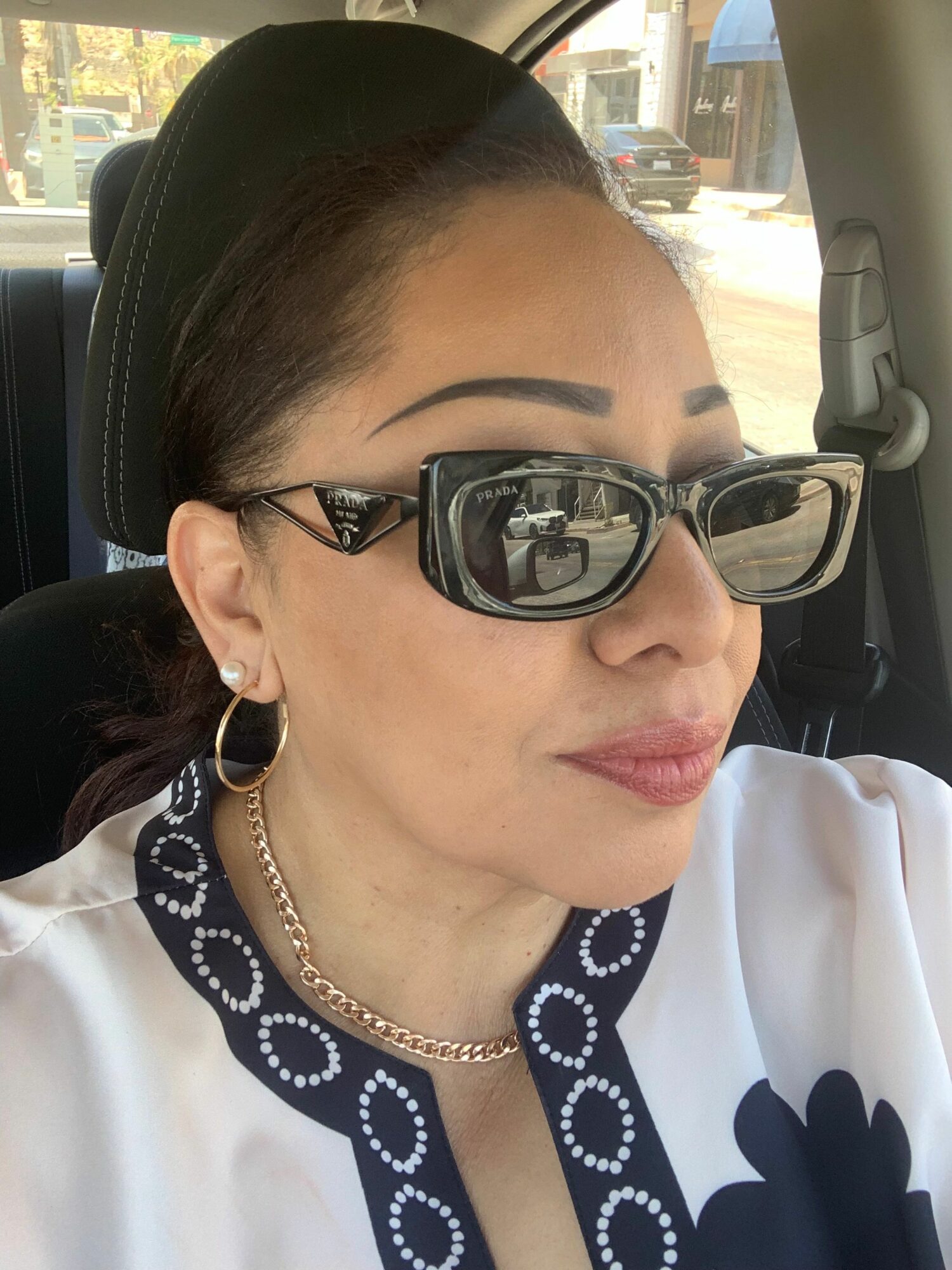 Woman with glasses, earrings, and necklace, wearing a white and black outfit, sitting in a vehicle, looking to the side.
