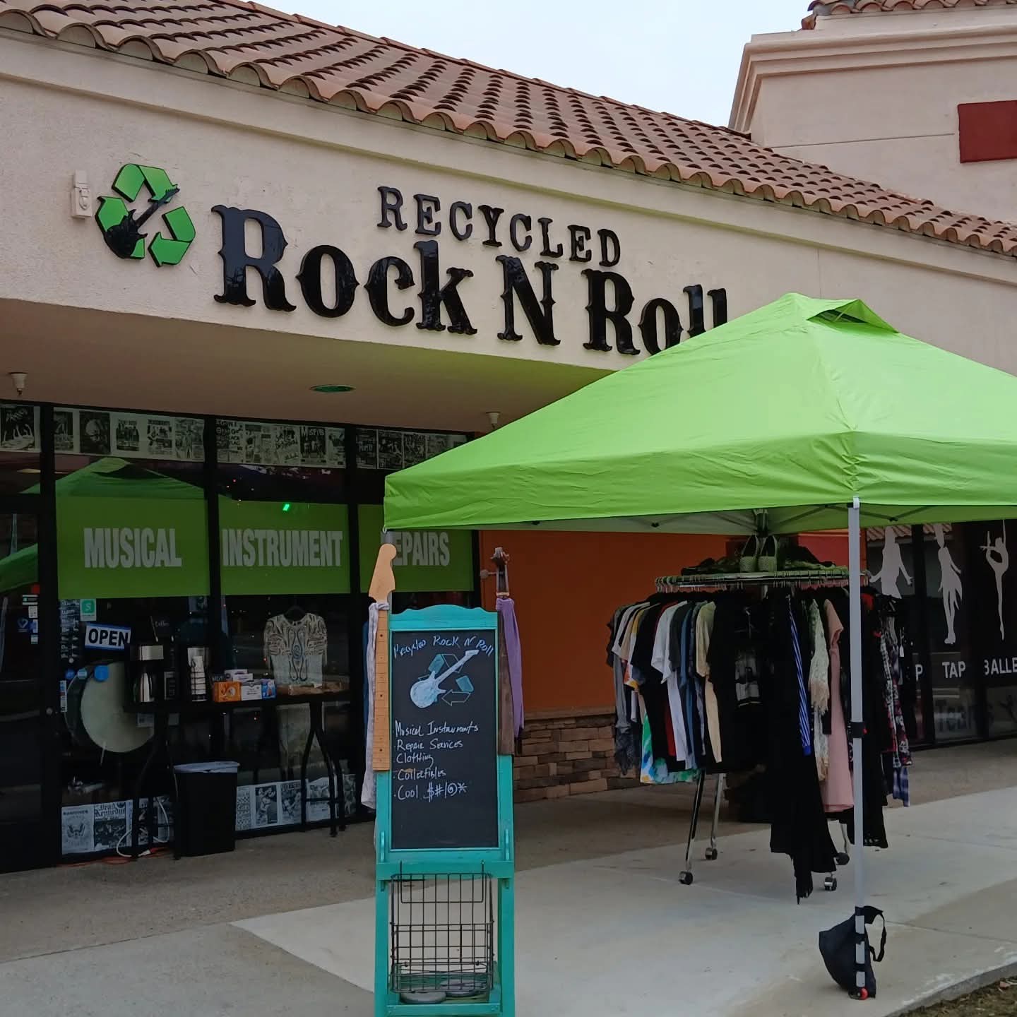 Storefront with a green canopy, clothing rack, and a sign for recycled Rock N Roll, with store windows and signage.