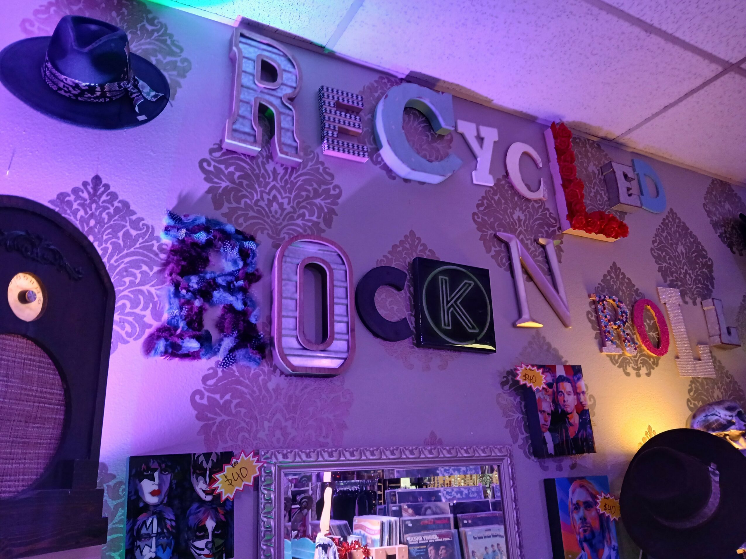 Decorative wall with large colorful letters spelling 'RECYCLING' and various decorations, including hats and flowers, in a room with purple lighting.