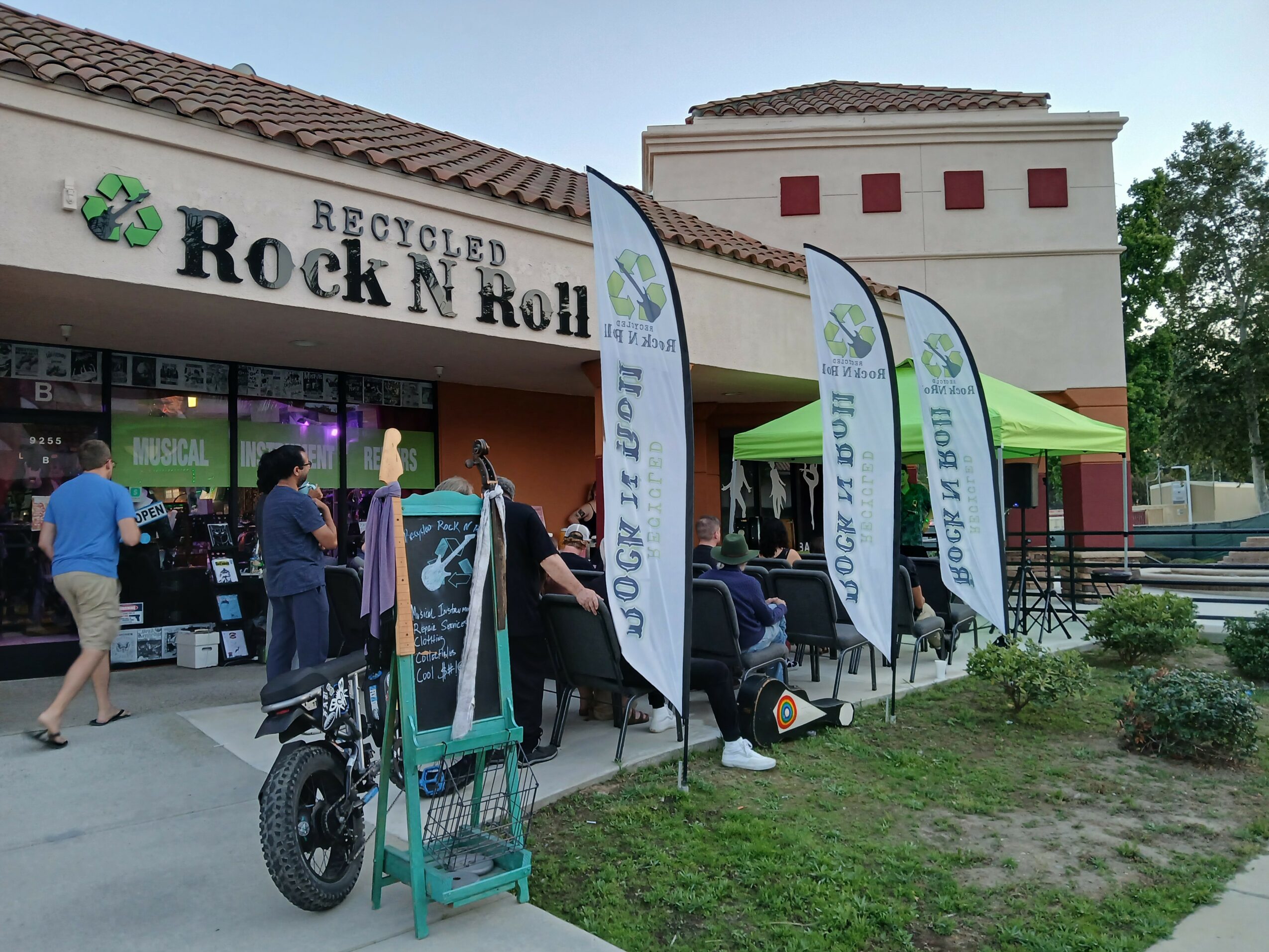 Outdoor event with flags, tables, and people near a building with a sign reading 'Recycled Rock & Roll'.