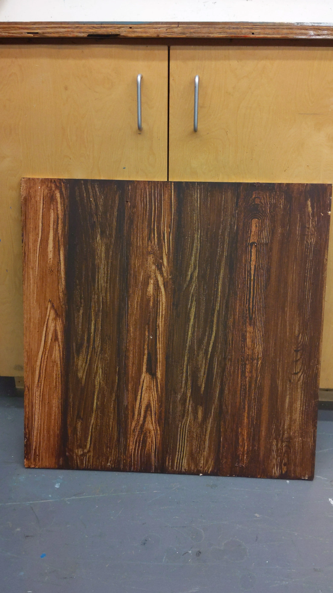 Wooden panel with a dark, textured surface leaning against a yellow cabinet with silver handles.