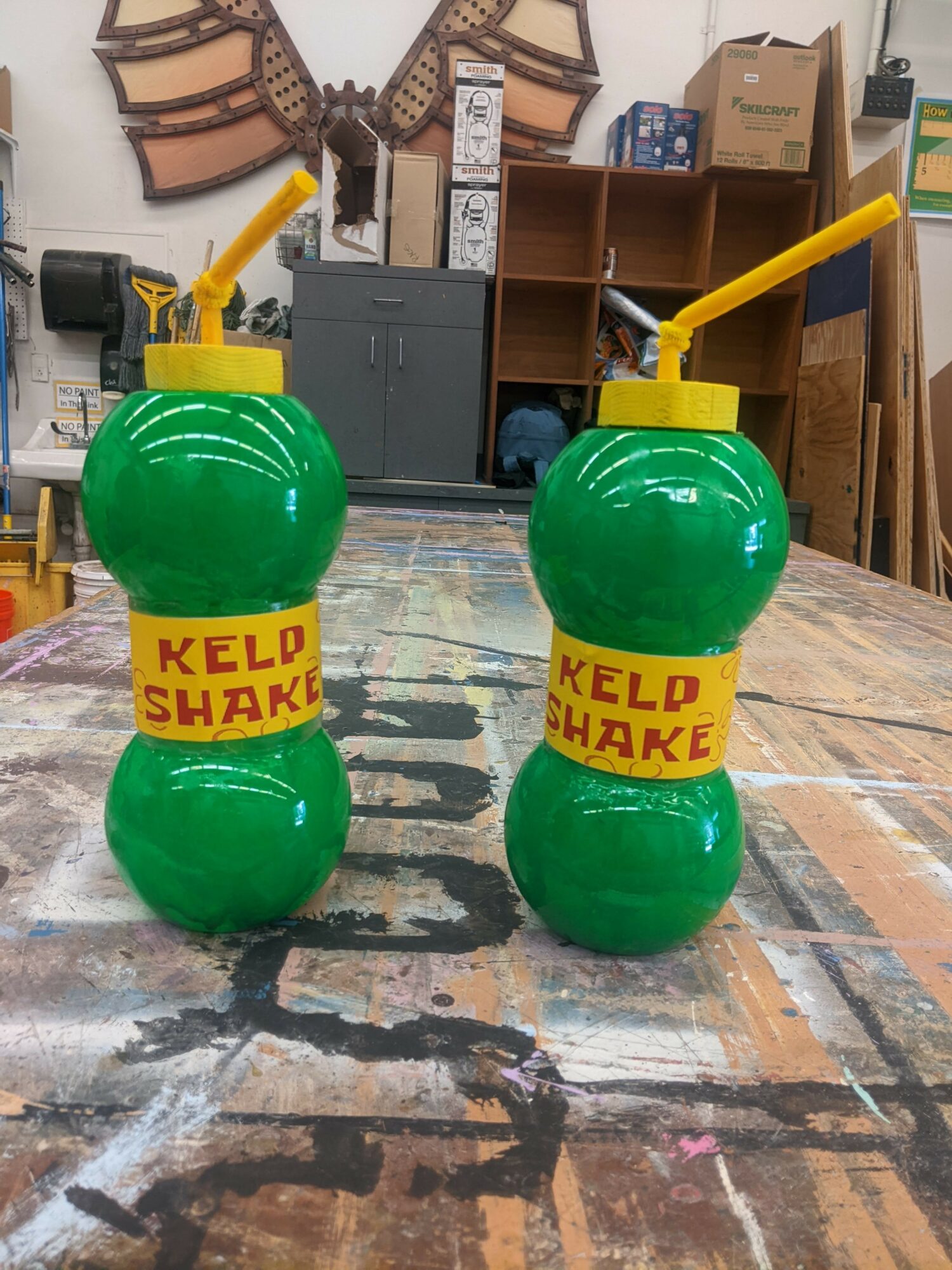 Two green plastic bottles with yellow labels reading 'KELD SHAKE' and yellow straws on a wooden table.