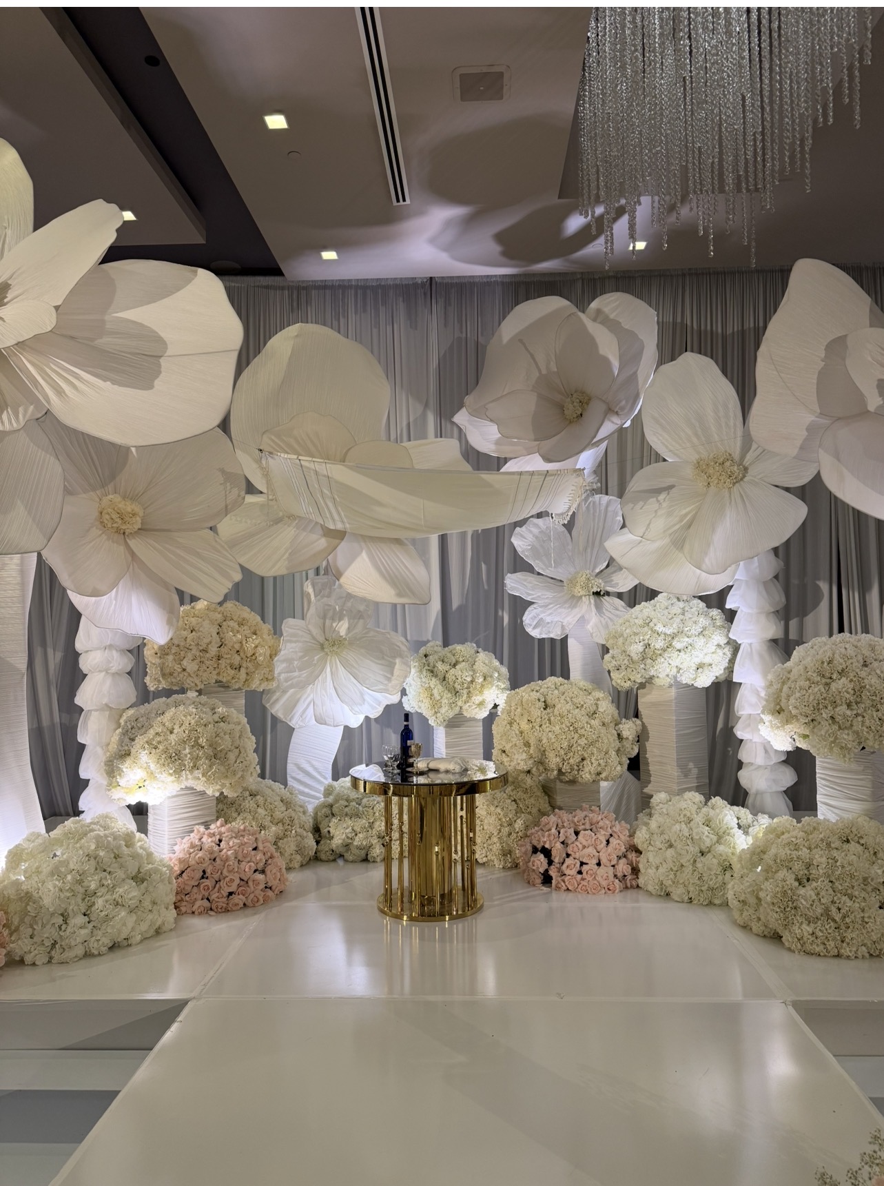 Elegant floral arrangement with white and pink flowers, a gold table, and a backdrop of curtains and hanging decorations.