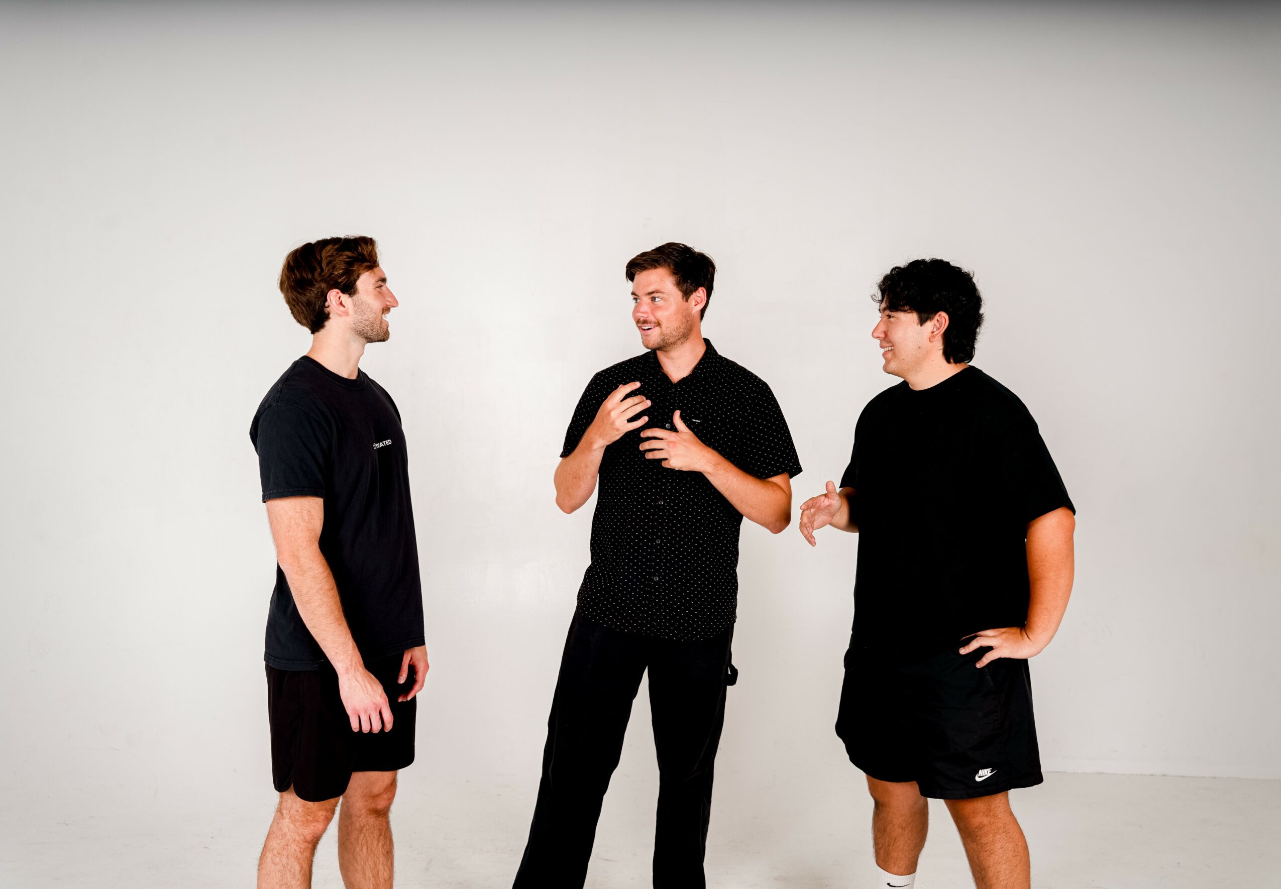 Three men standing and talking against a plain white background, engaged in conversation, with one gesturing with hands.