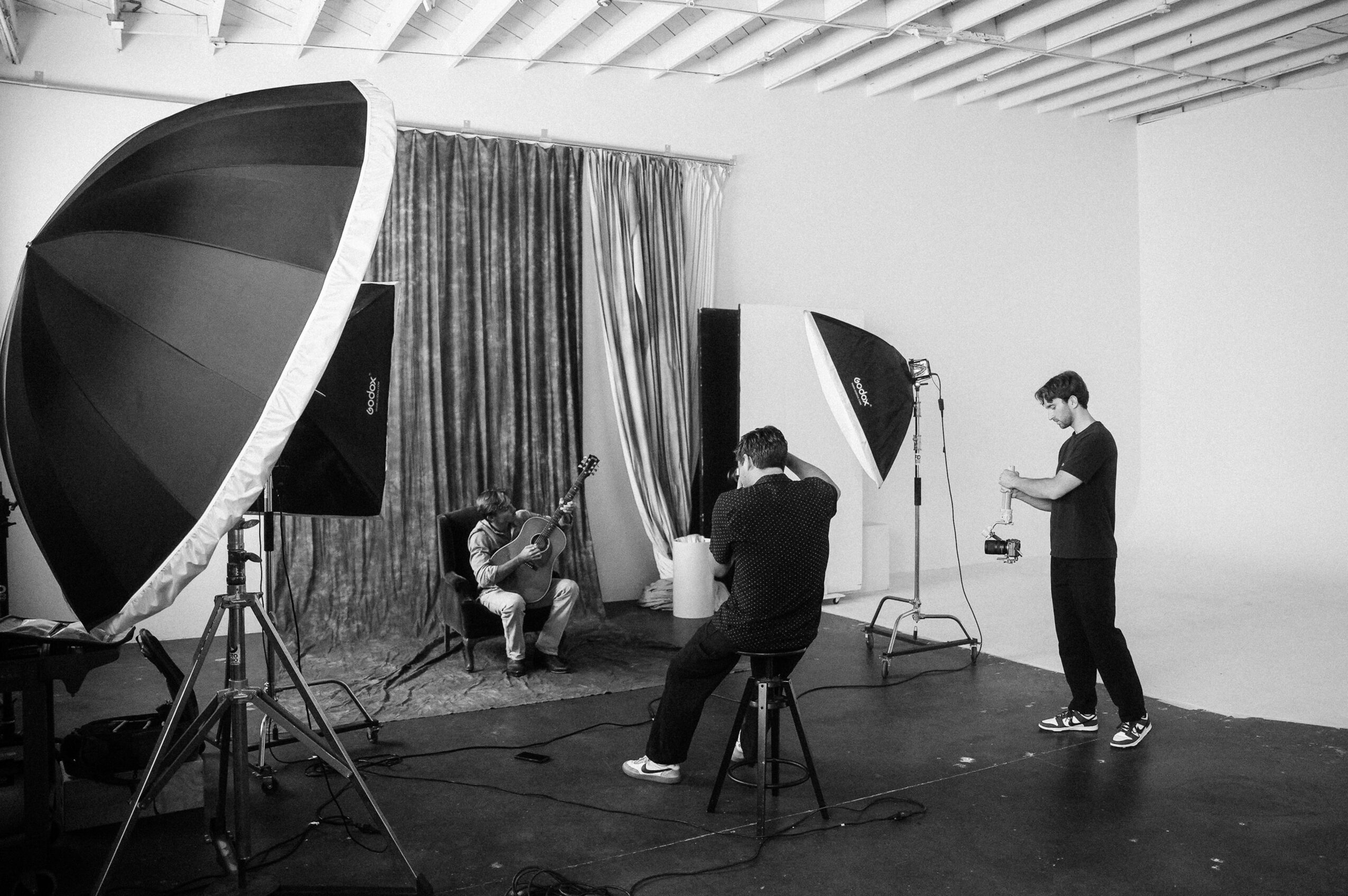 Photo of a photo shoot with a person sitting on a chair, surrounded by photography equipment and crew members, in a studio.