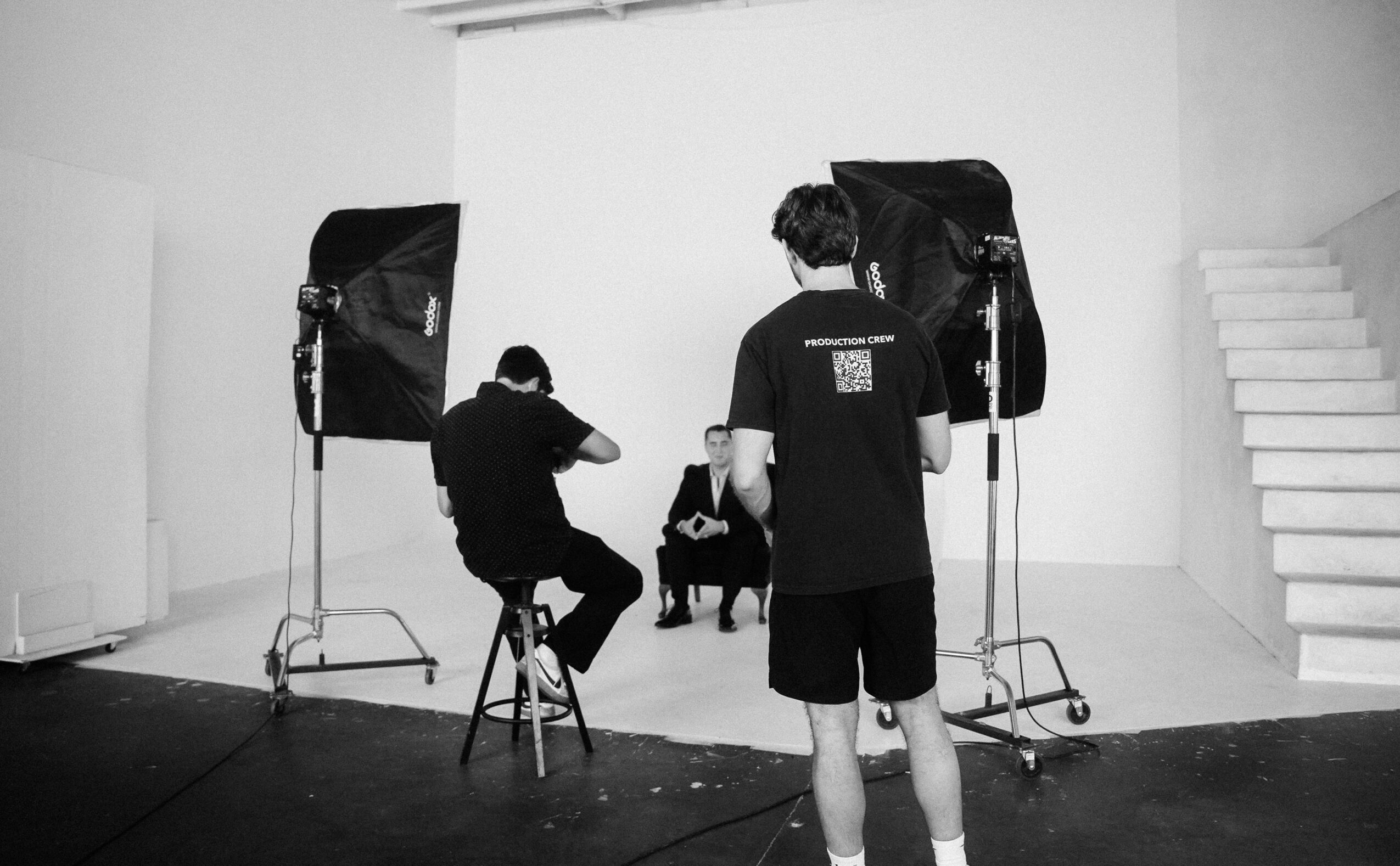 Two people in a photography studio with lighting equipment, one sitting and one standing, with a dog in the background.