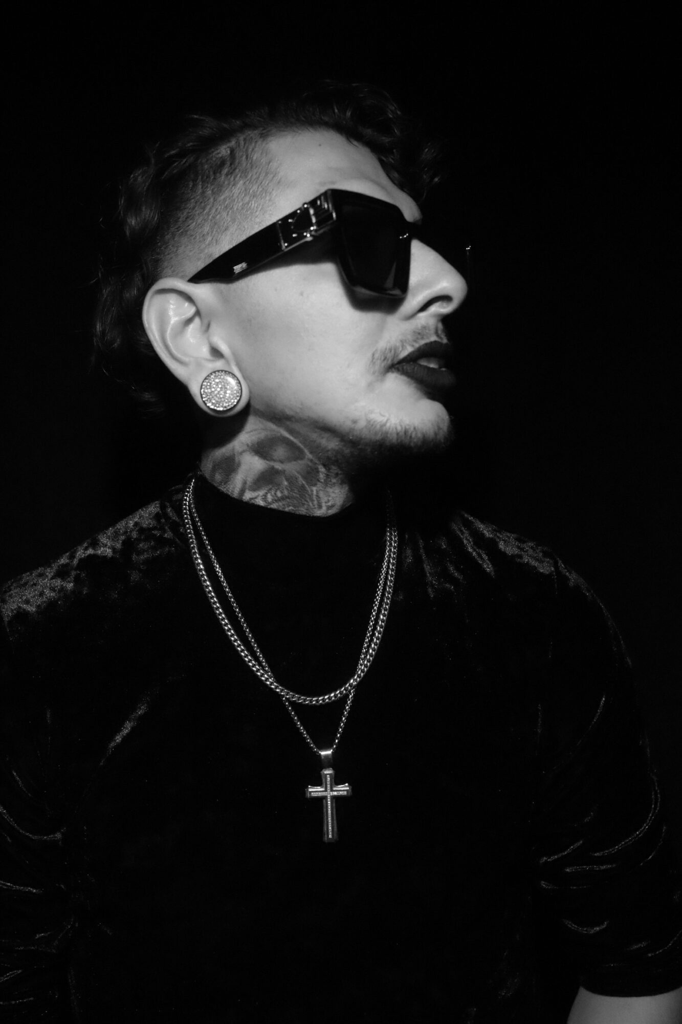 Person with short hair, sunglasses, earrings, and a cross necklace, facing sideways in a dark background.