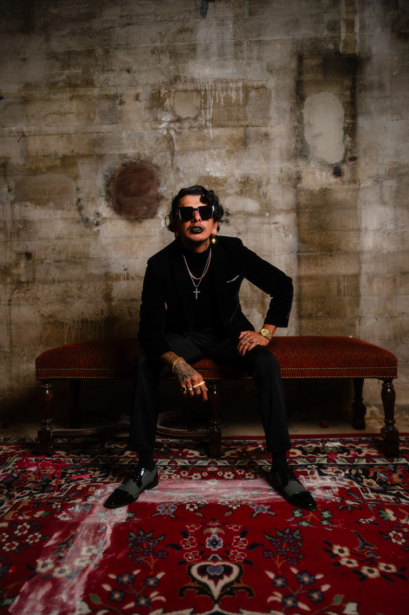 Person sitting on a bench in front of a textured wall, wearing dark clothing and sunglasses, with a patterned rug below.