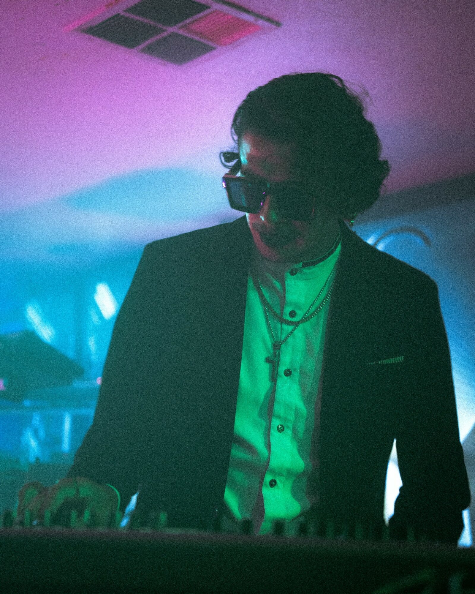 Person with curly hair and glasses wearing a suit and white shirt, playing keyboard in colorful lighting.