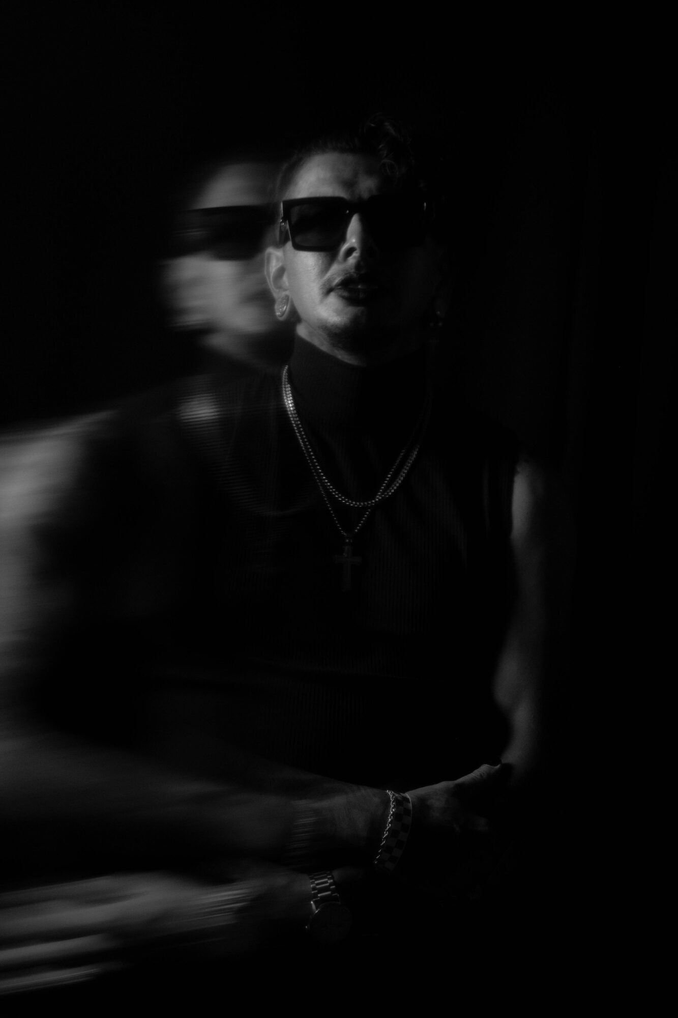Person wearing sunglasses and jewelry, with a blurred second figure in the background, in a dark setting.