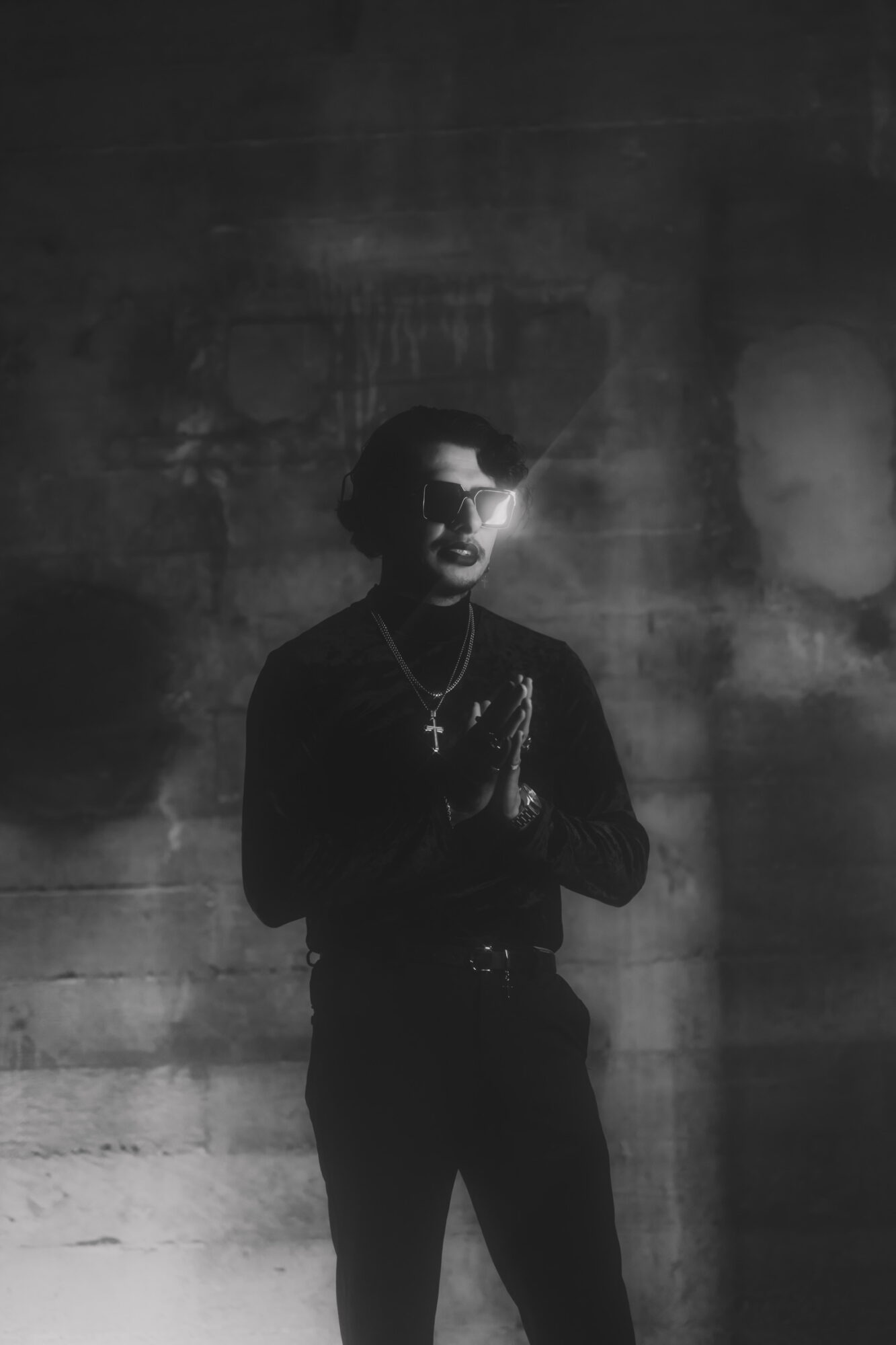 Person standing with hands together, wearing dark clothing and glasses, in front of a textured wall, in black and white.