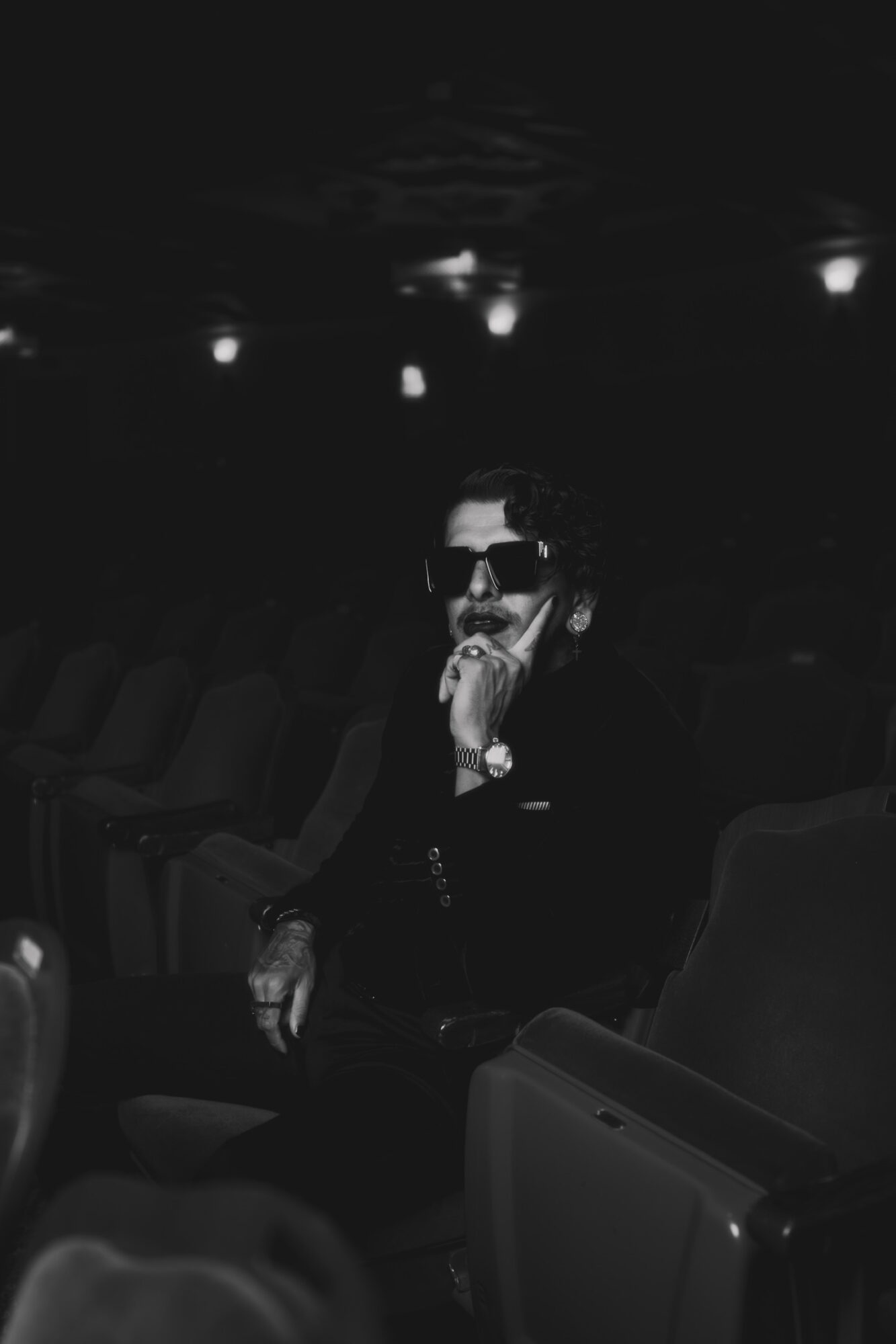 Person wearing sunglasses and dark clothing, sitting in a dimly lit theater, with hand near face, surrounded by empty seats.